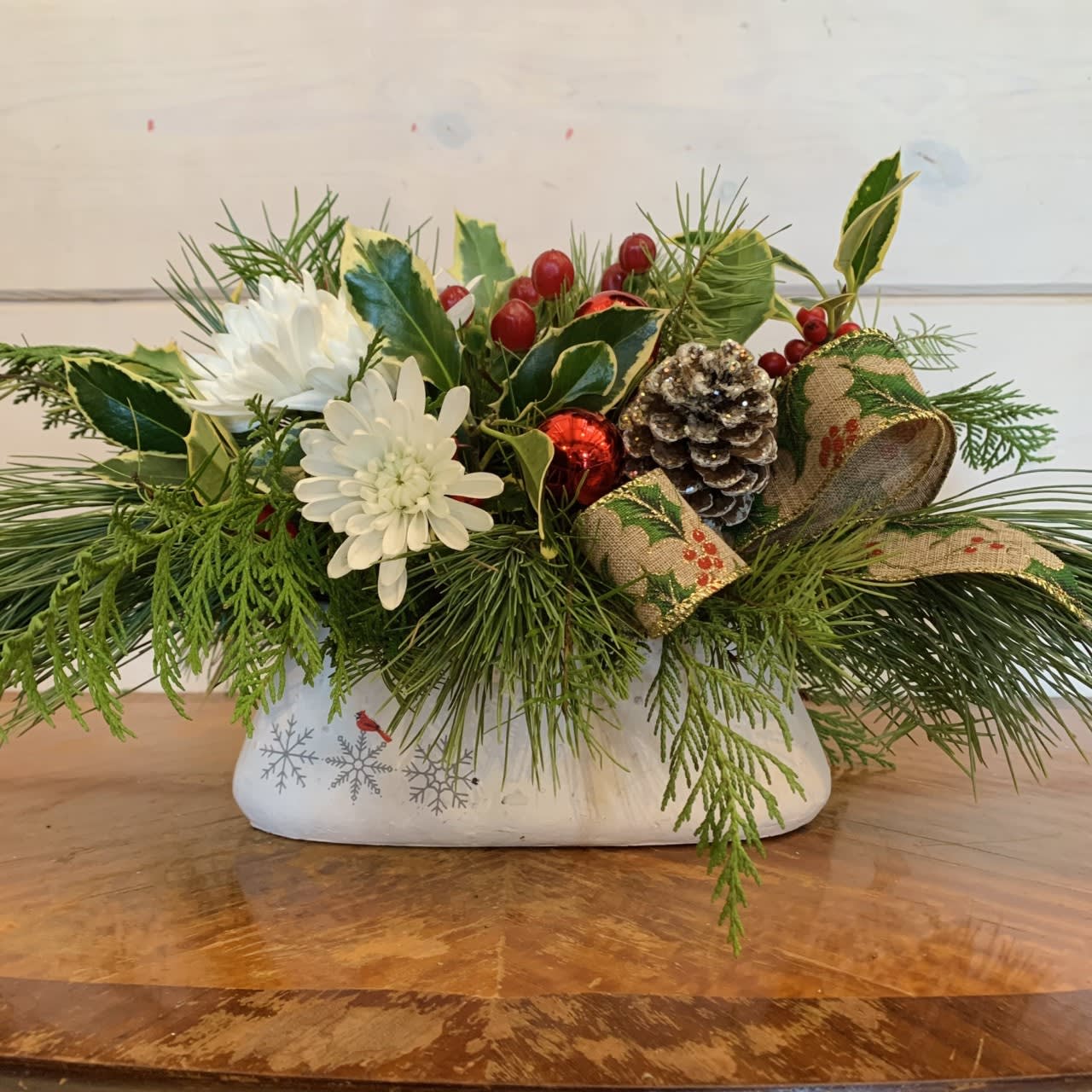 Holly Jolly Christmas  - Long lasting ingredients make up this sweet festive arrangement. winter evergreens, red hypericum berries, white chrysanthemums, arranged with lots of holly berries and pinecones bows and red ornament balls. Approximately 12in wide and 8 in tall. 