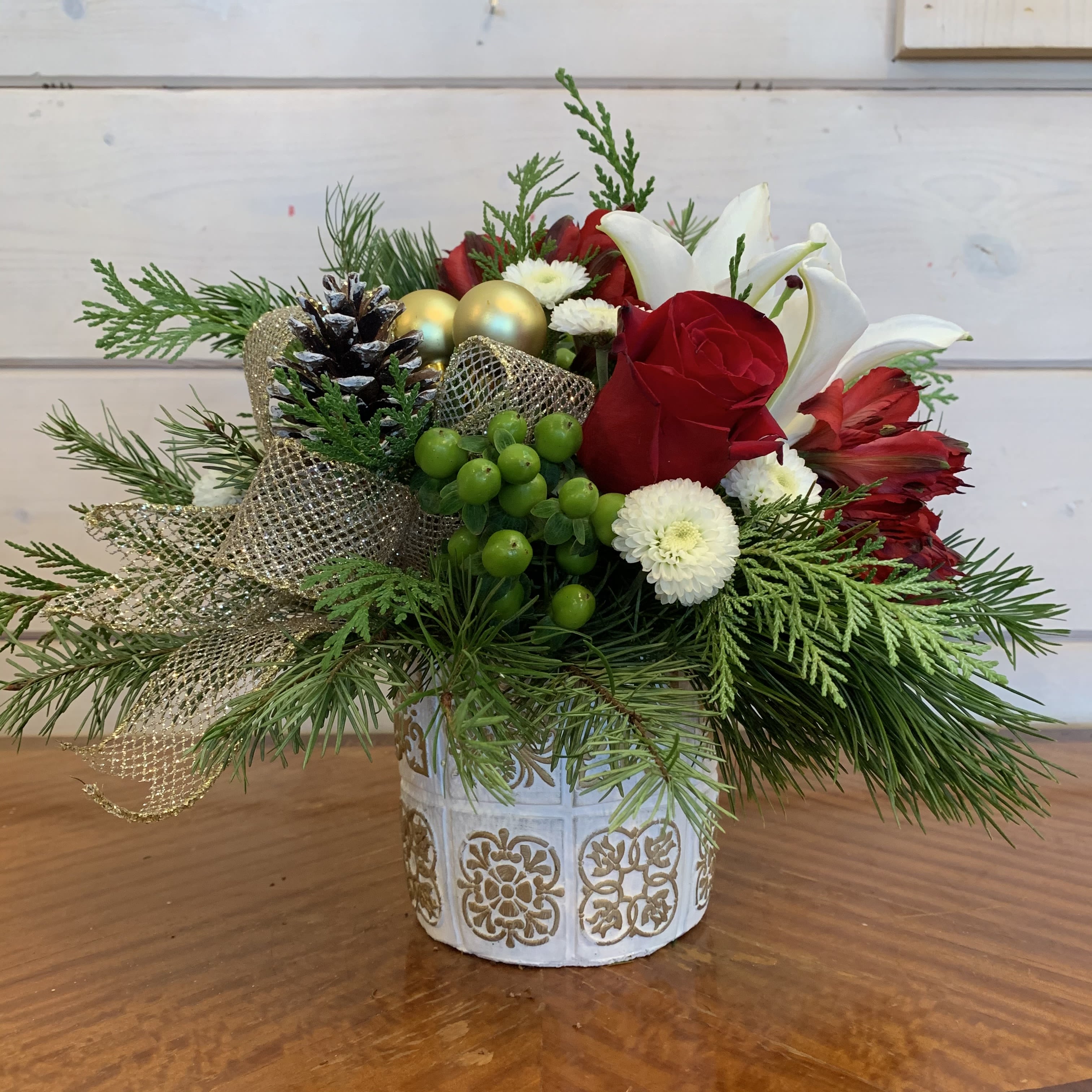 Making Spirits Bright - red roses and white lilies with green hypericum, white buttons, red alstroemeria, accented by snowy pinecones, gold ornament balls and a gold bow in a white pot with gold colored detailing. Approximately 12" wide by 12" high.
