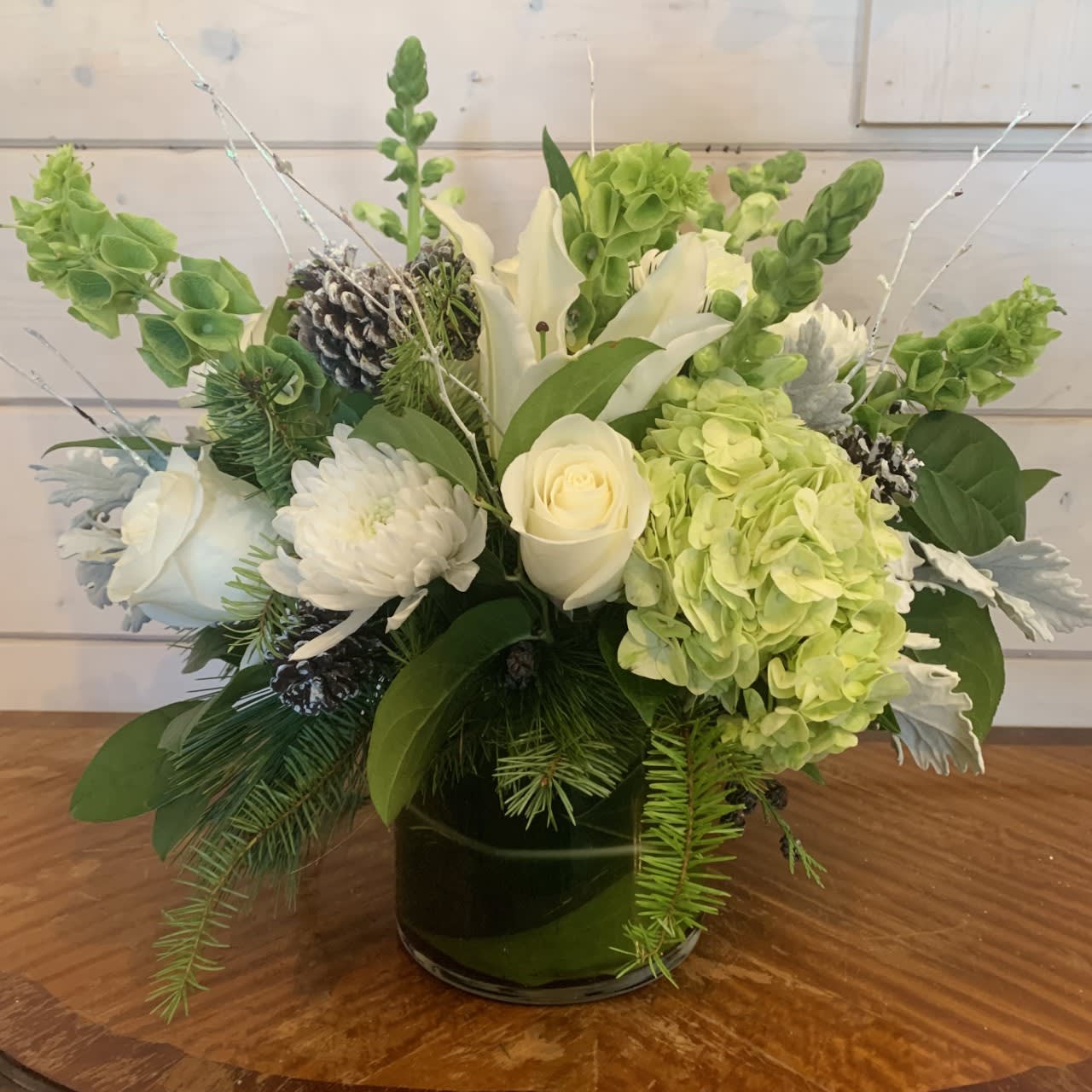 Peace and Joy to You Bouquet  - Elegant white and green arrangement in an extra large leaf lined gather vase. Includes a mix of roses, hydrangea, bells of Ireland, chrysanthemums, and dusty miller. Evergreens, white glitter branches and snowy pinecones make it festive for the season. Approximately 15in tall and 15in wide.