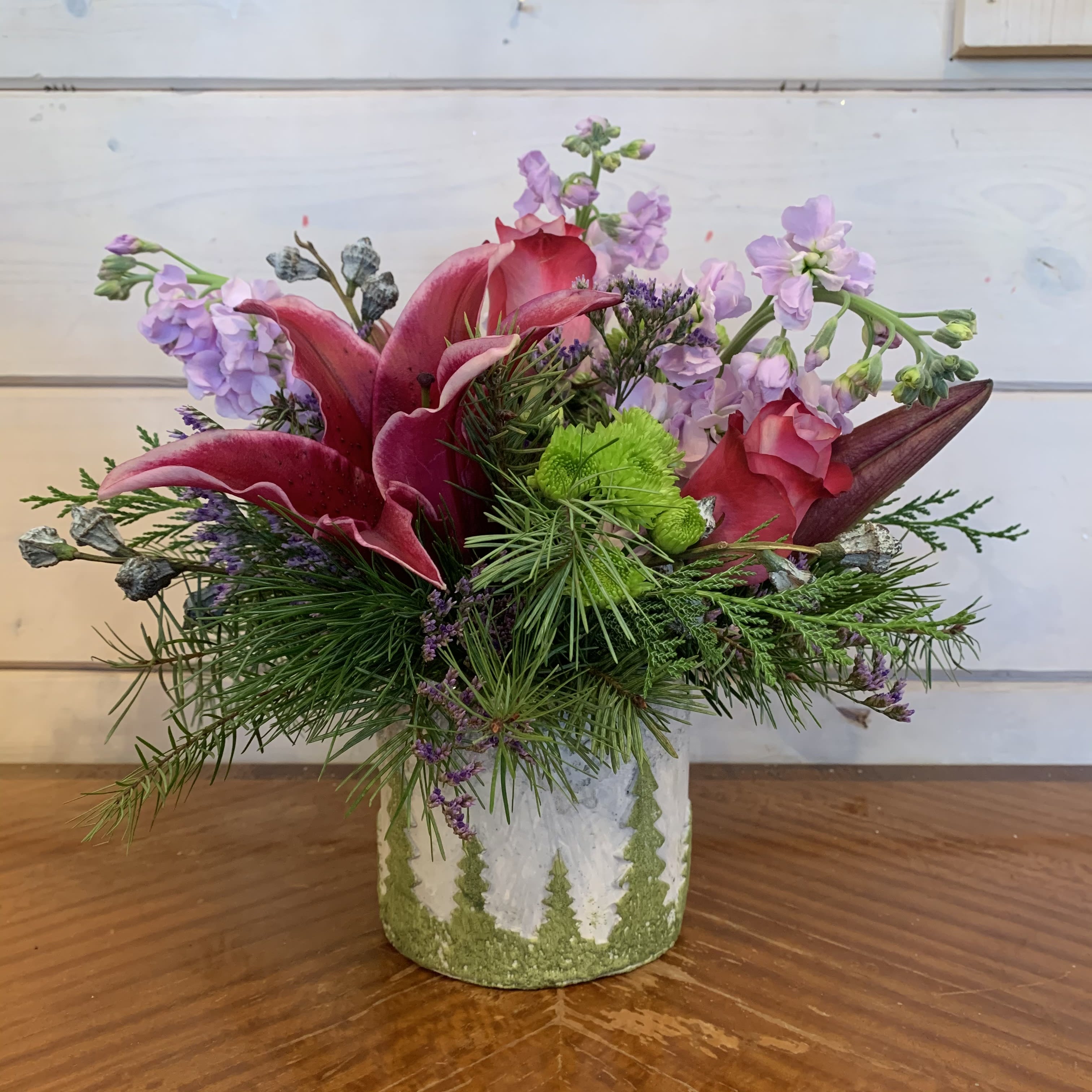 Starry Night - Lavender roses, pale lavender stock, burgundy lilies, green buttons, and purple misty arranged in a small textured ceramic vase with trees with seasonal evergreens and winter acorns. Approximately 10in tall and 10in wide.  