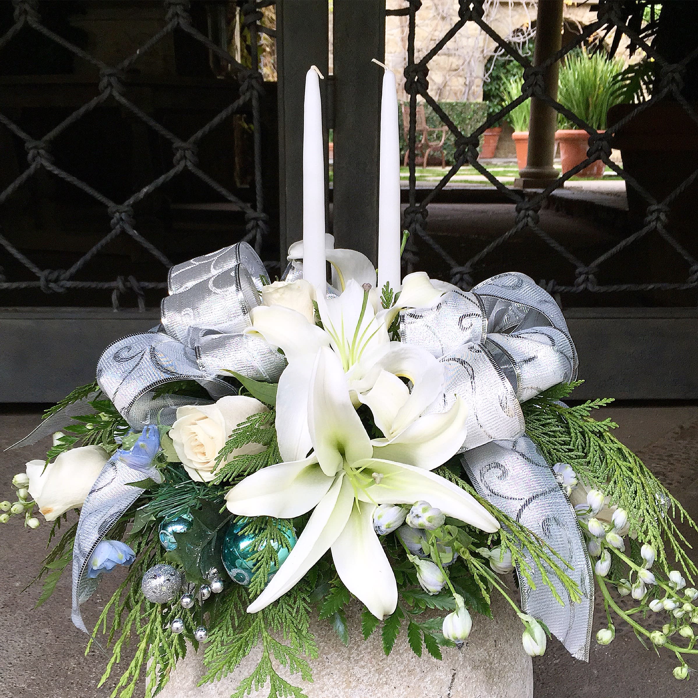 Snowy Holiday Centerpiece - Shimmering silver and blue with pure white candles to light up every table! This elegant bouquet designed with casablanca lilies, roses and delphinium will be the best gift to give for Christmas, Hannukah, Kwanzaa, New Year and all holidays you feel like celebrating.  Approximately 16" D