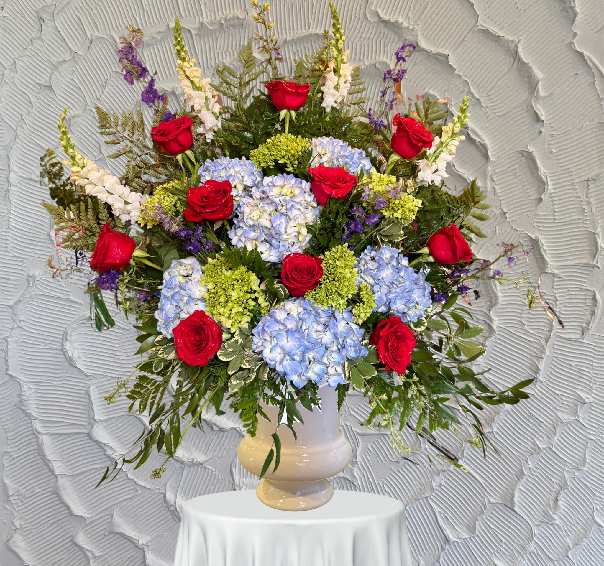 Benevolent Blooms - Pedestal Arrangement - This one-sided Pedestal Arrangement is made with Roses, Hydrangeas, Snap Dragons &amp; Delphinium.
