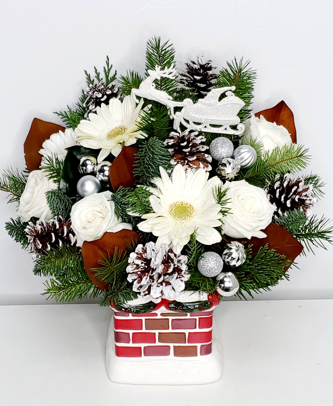 Meet Me at the Chimney Top - Custom Christmas  bouquet  uniquely designed with premium white roses, carnations, gerber daisies on a bed of Christmas greens and  magnolia leaves accented with silver ornaments and snow pinecones expertly curated.  In a ceramic Chimney Keepsake that would last long after the holiday is gone.  17"H by 12"W  DISCLAIMER:  Maybe designed in any color on hand. Please indicate color preferences under instructions.