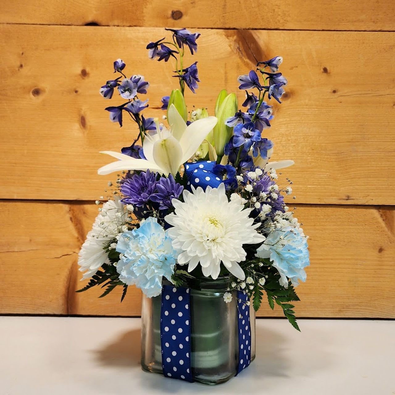 Polka Dot Prince - Welcome a new baby boy with this charming floral arrangement, thoughtfully presented in a clear glass cube and accented with a blue ribbon adorned with white polka dots. Standing approximately 16 inches high and 10 inches wide, this design is a delightful celebration of the little one's arrival.  The arrangement features a harmonious blend of blooms, including striking blue delphinium, which adds a bold and vibrant touch; soft carnations, offering a gentle, textured softness; cheerful cremones, with their bright and lively appearance; elegant lilies, contributing a touch of sophistication and fragrance; and delicate baby’s breath, which adds a light and airy finish.  Together, these flowers create a beautiful and celebratory display, perfectly suited for honoring a baby boy with a blend of elegance and joyful whimsy.