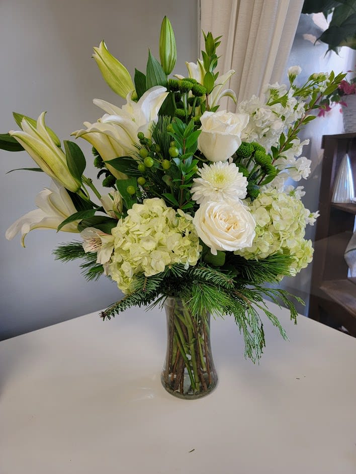 Winter White Blessings - A beautiful mix of white flowers with touches of green berries and mixed evergreens arranged beautifully in a gathering vase. Flowers include hydrangeas, lilies, alstromeria, stock, mums and roses.