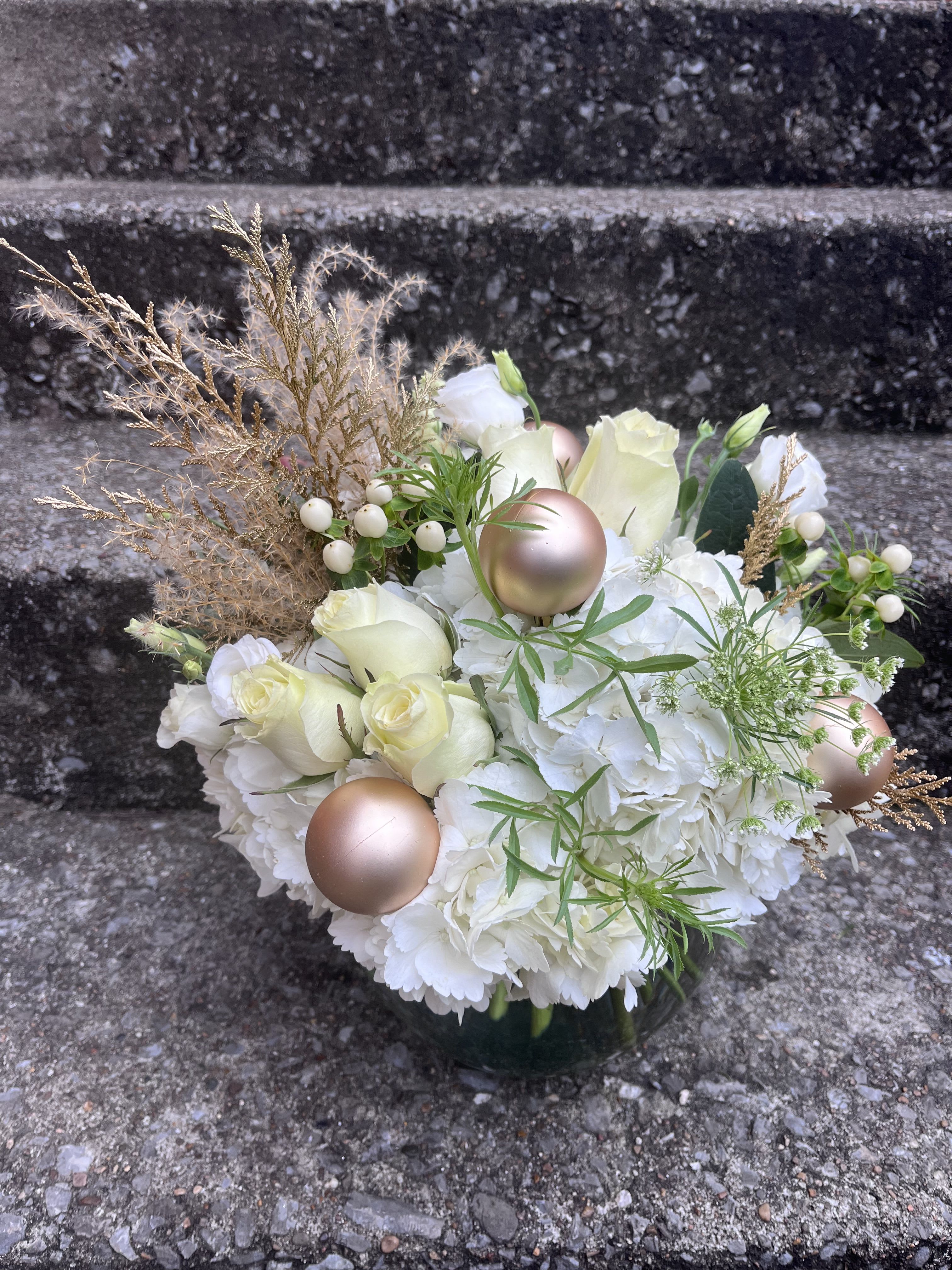 A Winters Glow - This arrangement has hydrangea, lisianthus, roses hypericum, queen Ann’s lace, Christmas bulbs and golden Christmas greens. The arrangement show above is the (deluxe size).