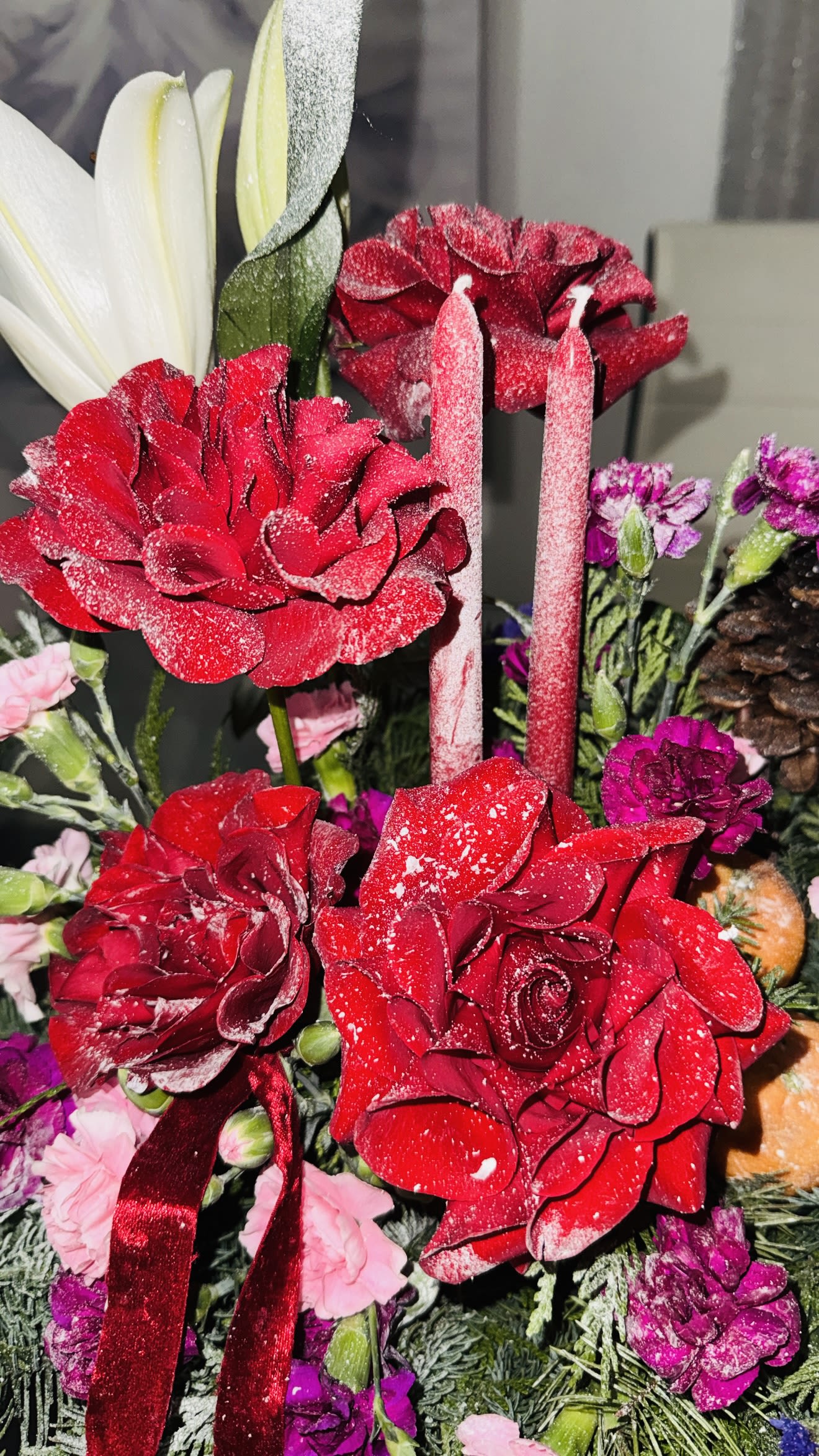 Winter Blessing Centerpiece - This centerpiece feels like a story whispered straight from a winter fairy tail- rich, festive, and full of symbolic warmth.  Deep red roses rise like glowing embers, while snowy lilies stand tall and serene, adding purity and elegance. Clusters of magenta and blush carnations bring softness and romance, balanced by the rustic charm of pinecones and evergreen branches brushed with a frosted finished.  Two red candles create a sense of ceremony- a moment of light, blessing, and celebration. The small tangerines tucked between the greens are a beautiful seasonal accent: a symbol of abundance, joy, and good fortune during the holidays. A velvet red ribbon drapes through the design, making the whole arrangement feel luxurious, festive, and full of heart. This is a centerpiece that brings the warmth of a winter home- glowing candles, the scent of pine, and the promise of a magic season. 