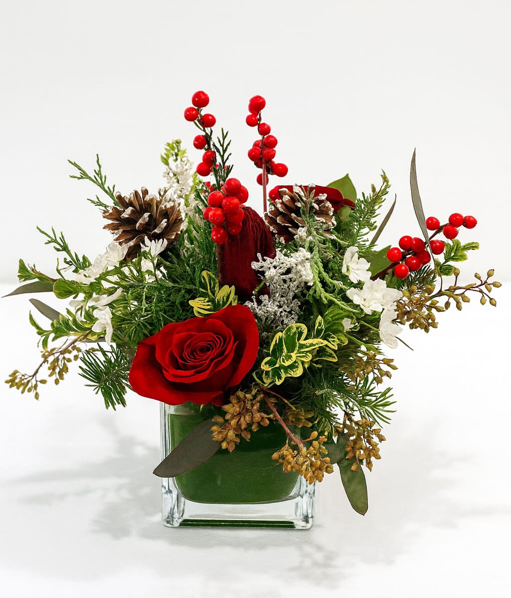 Winterberry Joy - A charming holiday arrangement featuring red roses, berries, red amaryllis, winter greens and pincones in a clear cube vase. Festive, cozy, and perfect for any space