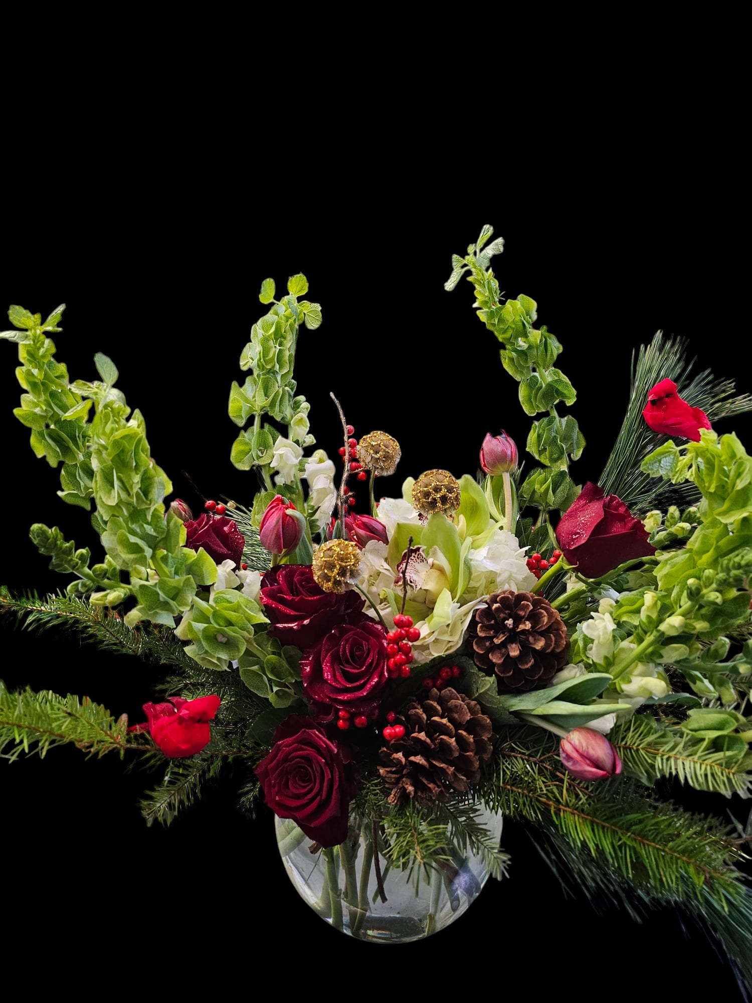 Traditional Christmas Elegance - Celebrate the season with a breathtaking display of classic holiday beauty. Our Traditional Christmas Elegance arrangement overflowers with lush seasonal evergreens, velvety burgundy Roses, and cheerful red Tulips, creating a luxurious arrangement full of warmth and charm. Clusters of festive berries and natural pinecones add rich texture, while elegant Bells of Ireland rise gracefully above the composition, giving height and a touch of winter magic.