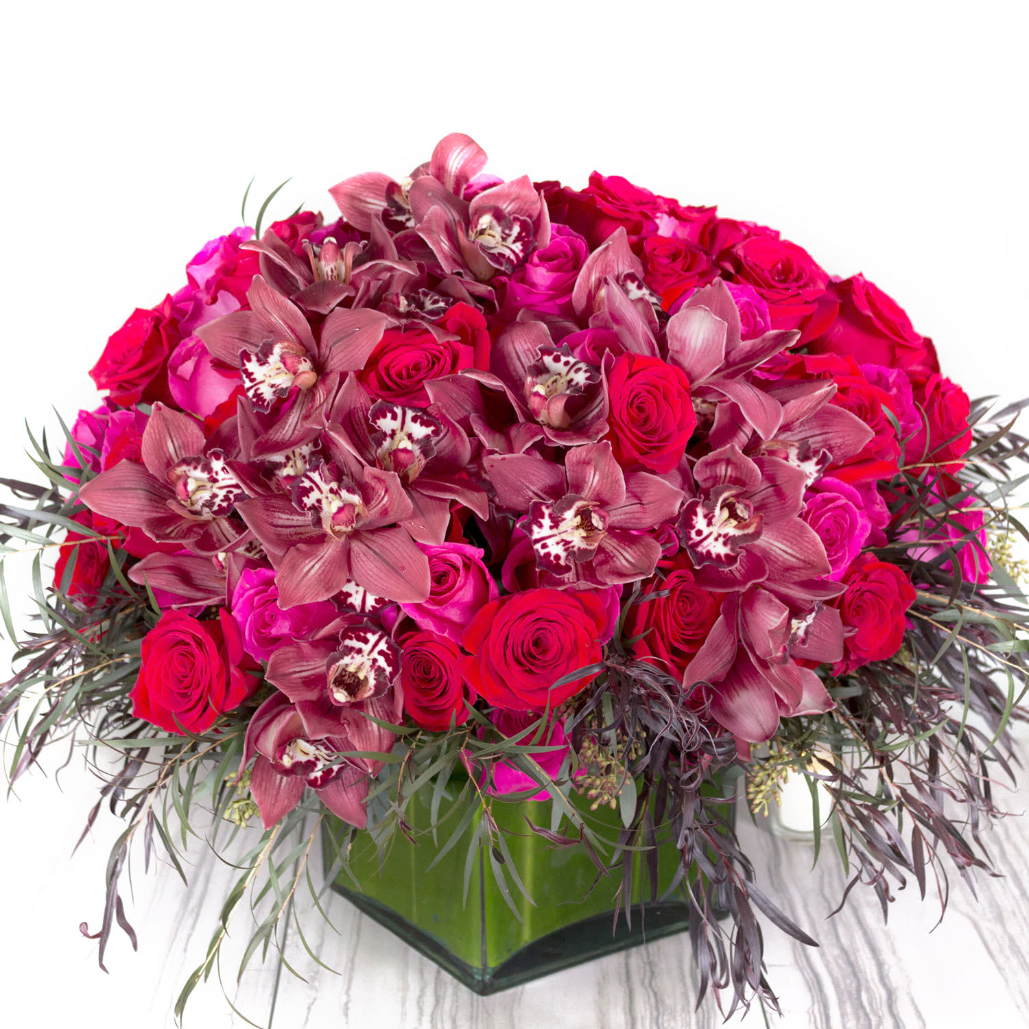 Red Reign (DB) - Prepare to be captivated by the sheer elegance and grandeur of our huge modern floral arrangement. This exceptional display features a magnificent combination of pink and white roses, perfectly complemented by the dark allure of cymbidium orchids. All of this beauty is impeccably designed within a large leaf-lined glass cylinder vase.
