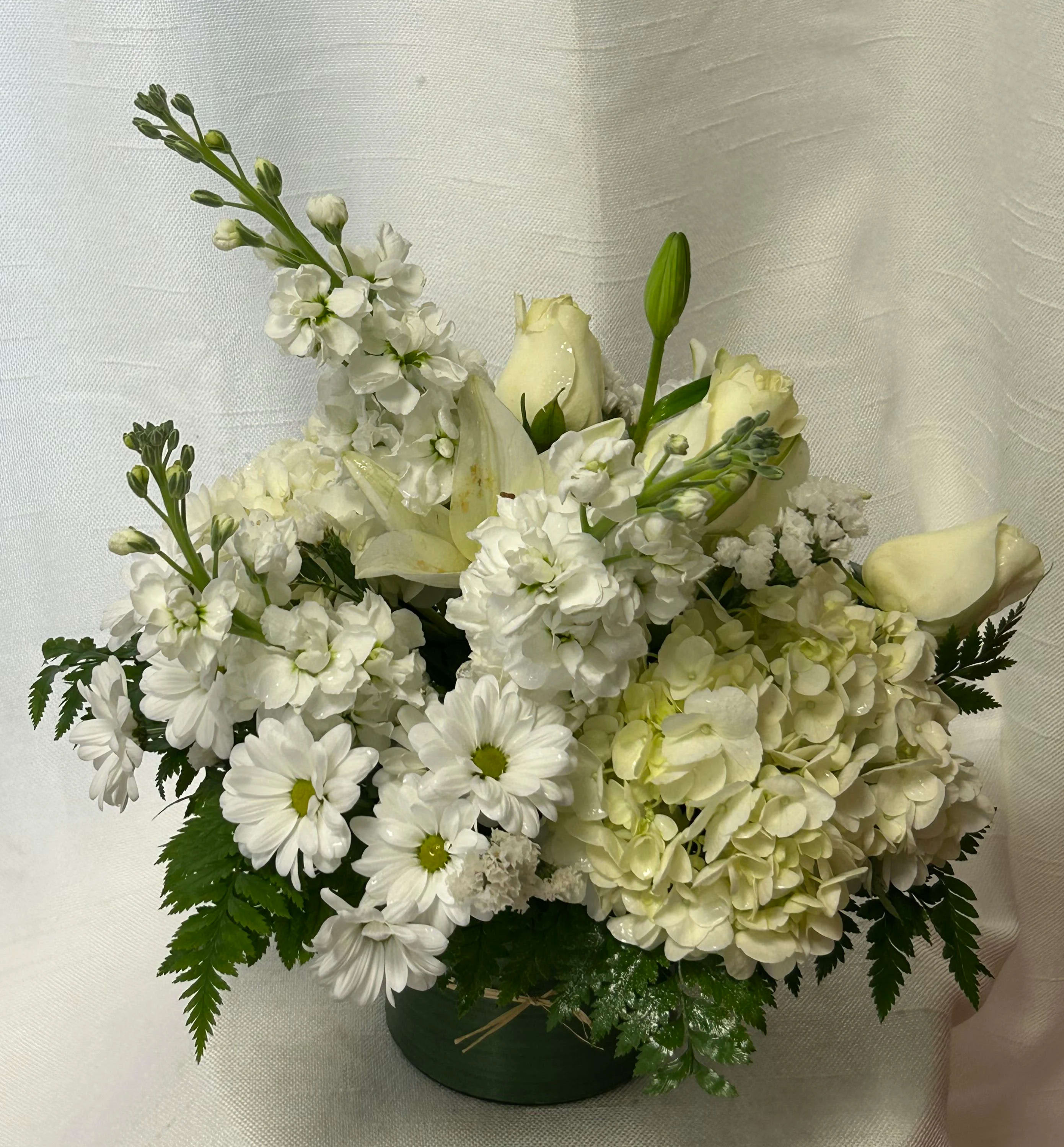 Cream De La Cream (DB) - Prepare to be captivated by the sophisticated allure of our all-white modern flower arrangement. This stunning display features a harmonious combination of hydrangeas, stock roses, daisies, and assorted greenery, all impeccably arranged within a leaf-lined glass cylinder vase.