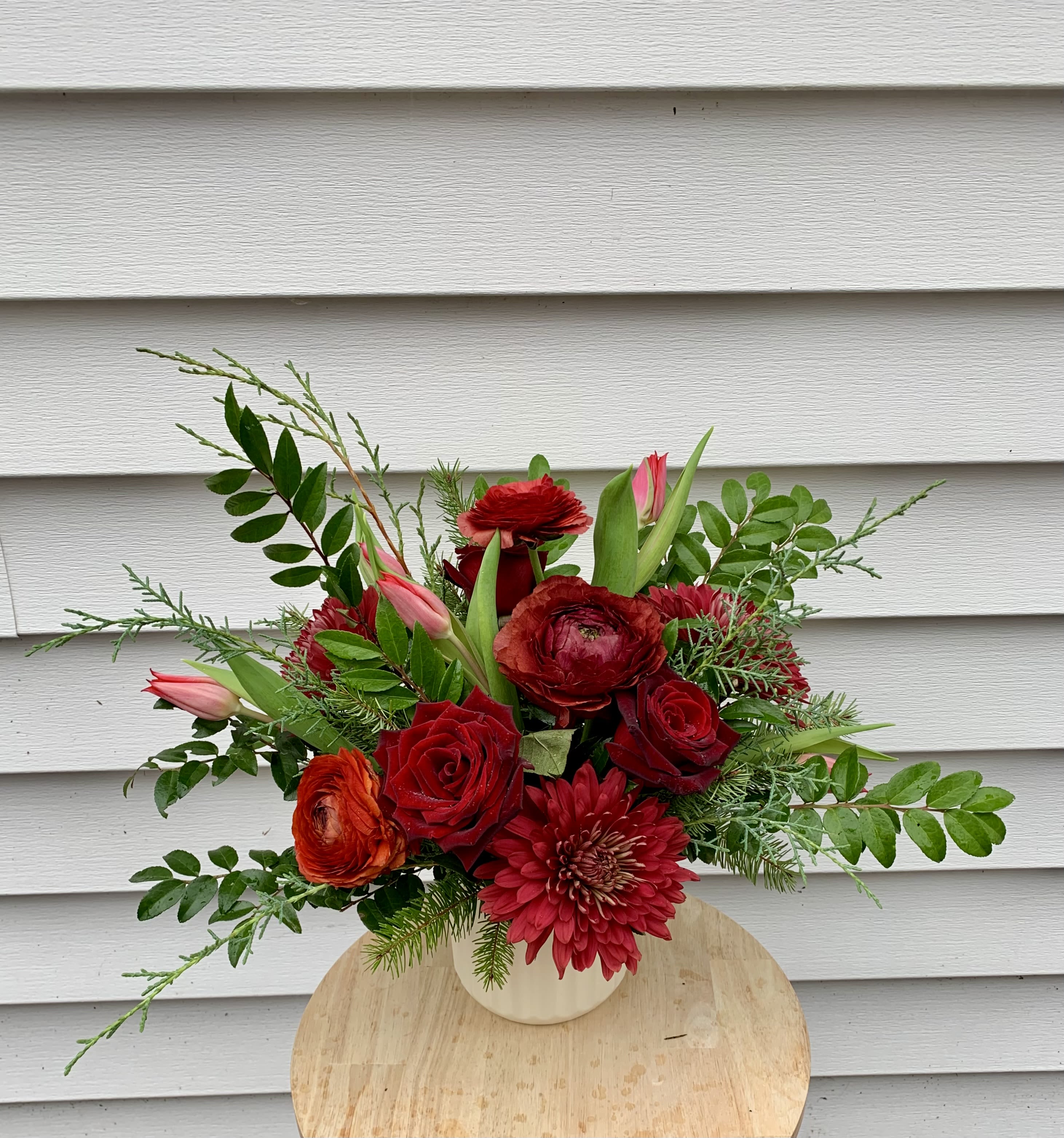 Christmas Classic - Bring the warmth of the season indoors with this festive red-and-green arrangement. Vibrant red blooms are paired with fragrant pine, cedar, and huckleberry evergreens, creating a lush and timeless holiday display. Designed in a clean, 5-inch white ceramic vase, this piece adds the perfect touch of Christmas elegance to any table, countertop, or gift exchange. Seasonal, fragrant, and full of cheer — a beautiful way to celebrate the holidays