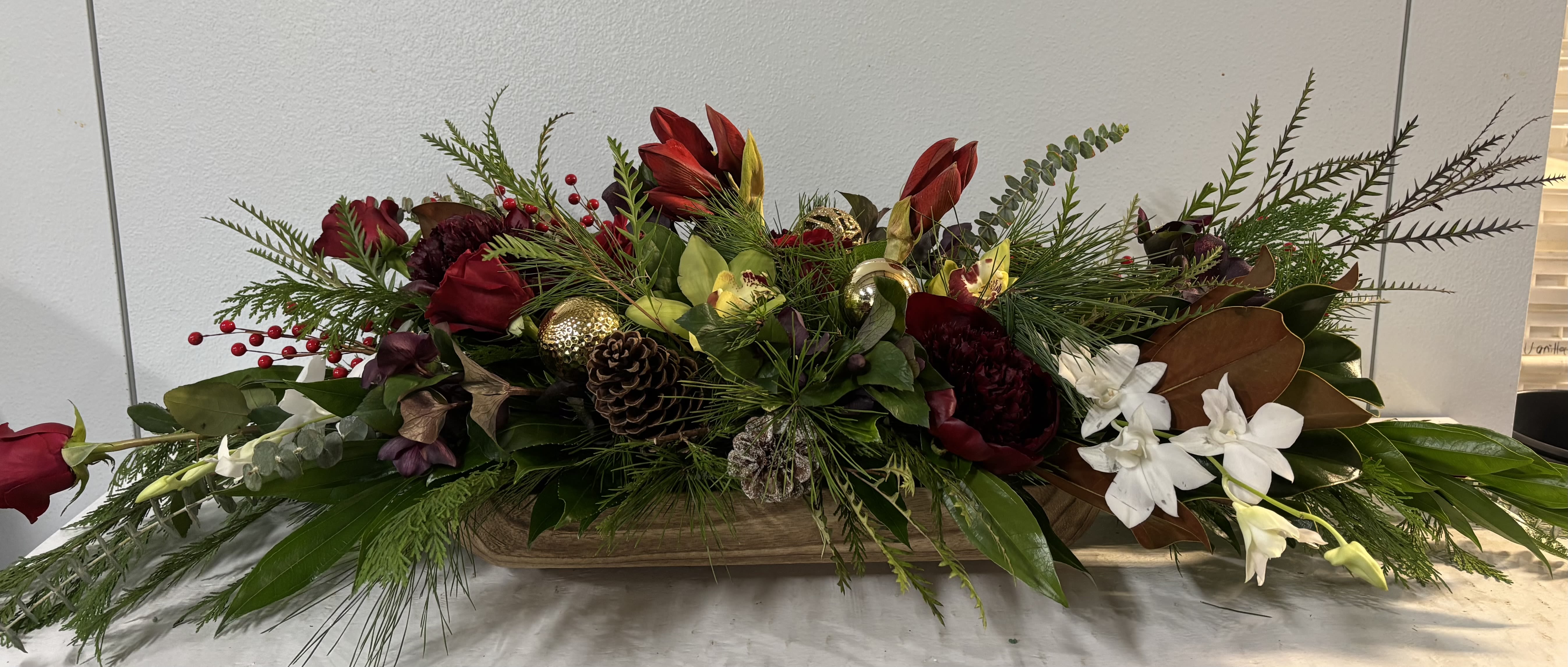 Majestic Christmas - Extraordinary centerpiece in wooden Dough Bowl. Measures 48" long x 16 wide. *You can specify other measurements to accommodate your table.*  This is a traditional blend of Stunning holiday greens including magnolia leaves, pine cones and berries. Flowers are Amaryllis, roses, peonies, dendrobium and cymbidium orchids and helleborus. And of course some bling....this is stunning!