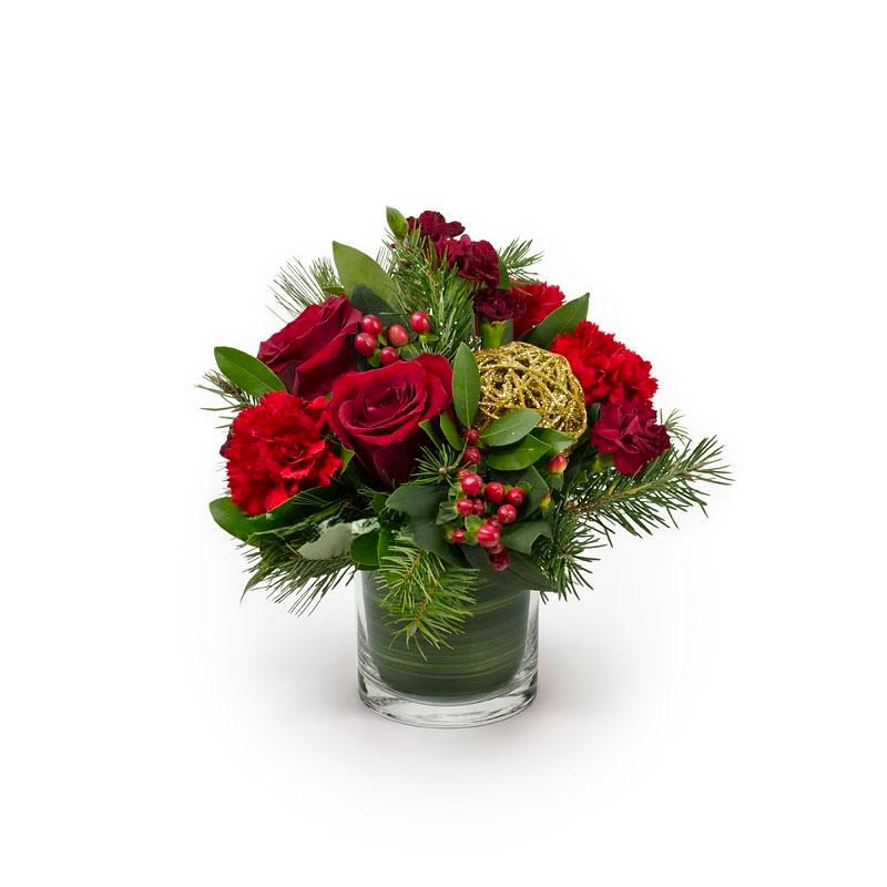 Holiday Joy - Red roses, carnations, holiday berries seasonal greens with  gold twig sphere accent. Arranged in a 4" cylinder vase lined with green lea ribbon. 