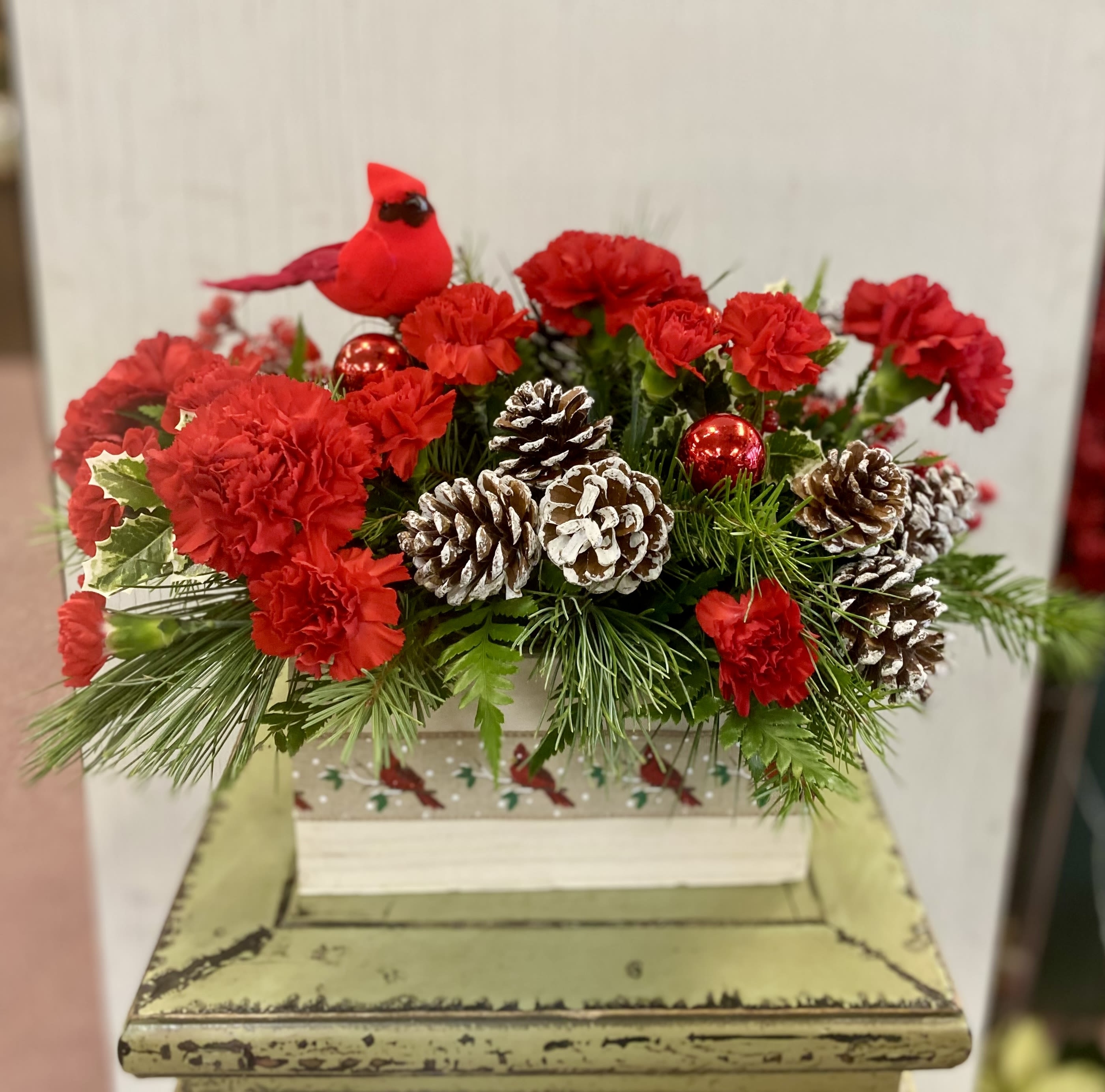 The Christmas Cardinal Arrangement - A bright and cheery cardinal sits atop this long lasting floral arrangement in vibrant holiday tones. ***CONTAINER WILL VARY, but all will be festive and cheery.
