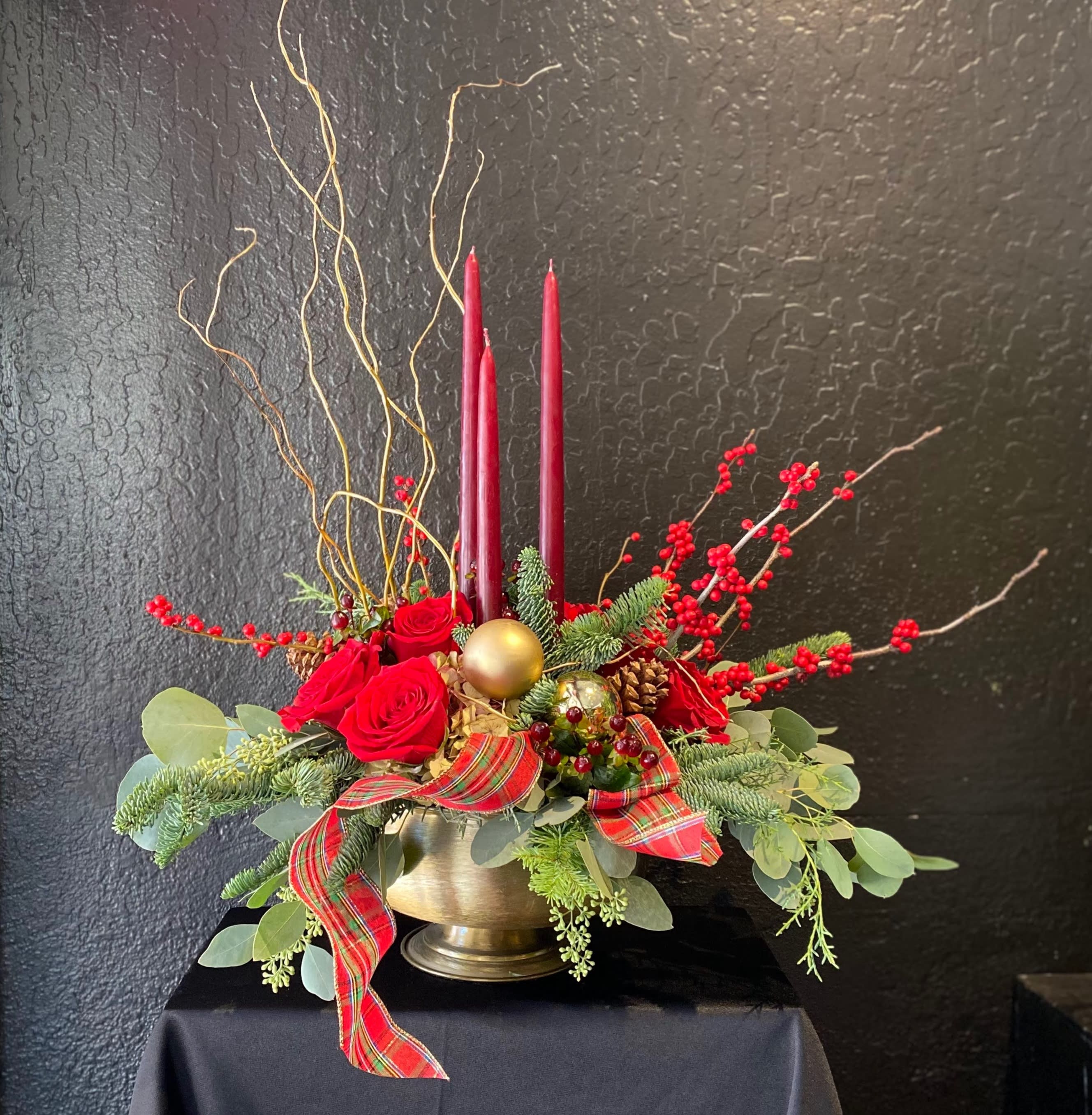 Warm Winter Song - An ode to classic winter style designed in a gorgeous golden-footed compote.  Arrangement includes 3 tapered candles, tufts of ribbon splaying elegantly down, and seasonal staples like ilex berries, roses, pine cones, and winter greens.
