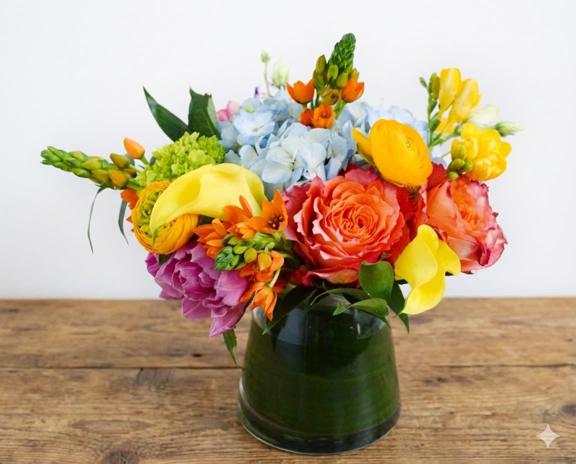 Warm Greetings - Whether for yourself or as a gift, our Warm Greetings vase arrangement will leave you feeling loved and happy. This arrangement combines a few of the classic flowers you know, but in a colorful design, along with a few others we love! Standard size pictured. Larger sizes available. Flowers and vase will vary based on availability. Sweetly scented yellow freesia, blue hydrangeas, yellow ranunculus, orange free spirit garden roses, yellow or white calla lilies and assorted fillers and greens. 