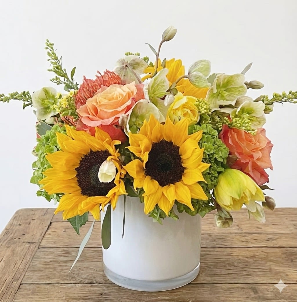 Picnic  - This arrangement include orange free spirit roses, sunflowers, hellebore, hydrangea, daffodils and assorted greens and fillers. It is presented in a white glass vase.  