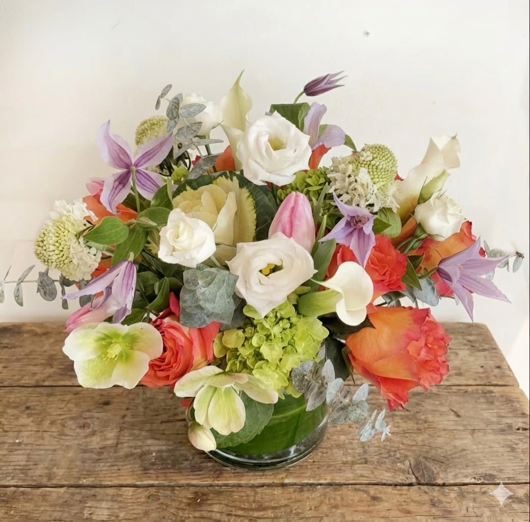 Garden Muse - Whimsical, fresh, and full of movement — the Garden Muse arrangement brings the essence of a blooming spring garden indoors. This lush design features a joyful mix of seasonal blooms including coral and blush roses, soft pink tulips, creamy lisianthus, white calla lilies, green hydrangeas, and delicate lavender clematis. Hints of eucalyptus and textured filler complete the look with airy elegance.  Presented in a classic clear glass vase, this bouquet is ideal for birthdays, thank-you gestures, or simply brightening your space with nature’s beauty.  Details: 	•	Premium mix of roses, tulips, lisianthus, clematis, hydrangea, and more 	•	Approx. size: 9” H x 10” W 	•	Designed in a clear reusable glass vase 	•	Includes a complimentary message card  Care Instructions: Keep in a cool spot out of direct sunlight. Change water daily and trim stems slightly every few days to extend freshness.