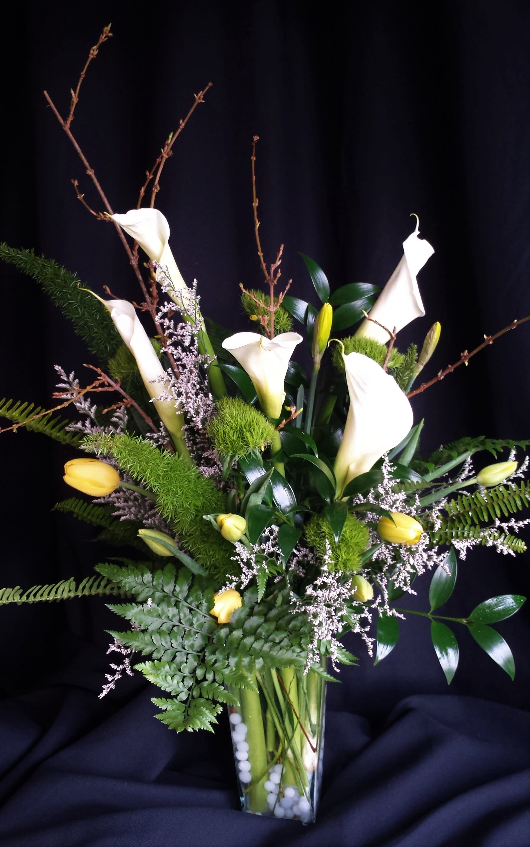 Winter fern and bulb arrangement - Tall bouquet of winter bulb flowers such as tulips daffodil and calla lilies as shown or similar flowers. Lush greenery and a rectangular vase with vase filler decor.