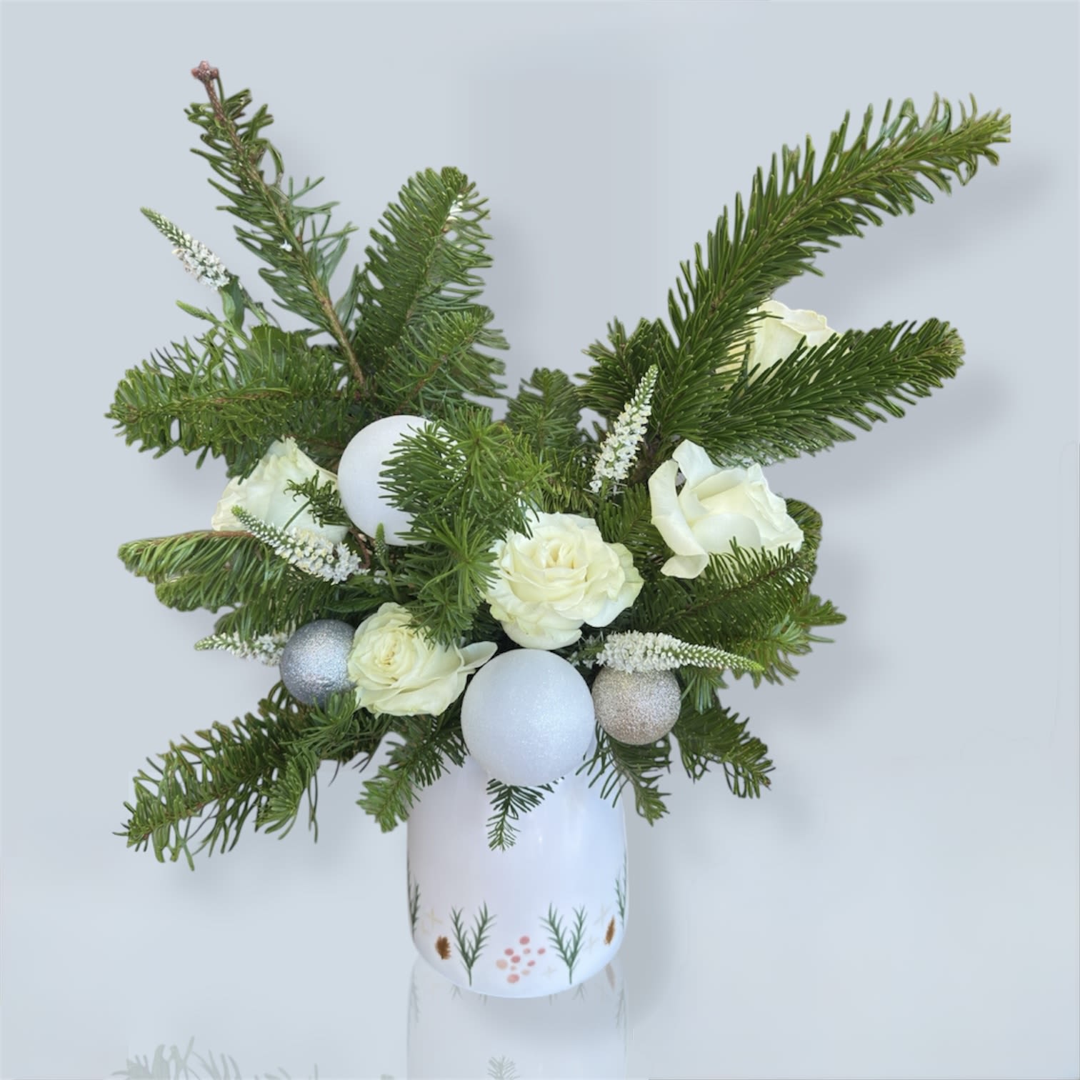 White Harmony - White Harmony is a fresh and elegant arrangement featuring white roses paired with natural pine branches to create a clean, modern piece perfect for the season. Subtle pearl-toned ornaments add a gentle touch of sparkle while keeping the design natural and refined. The white ceramic vase with botanical details gives it a soft, handcrafted finish. A bright, serene, and stylish arrangement that elevates any space.