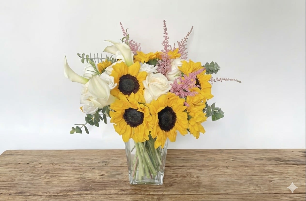 Stephanie  - Named after one of our lovely customers who helped create this design, the Stephanie comes in a tall, modern clear vase, making it the perfect centerpiece for an open space like a kitchen island. This vase arrangement includes bright yellow sunflowers, white roses, white cala lilies, and one of our favorite feather-like flowers, pink Astilbe. Flowers and vase will vary based on availability.