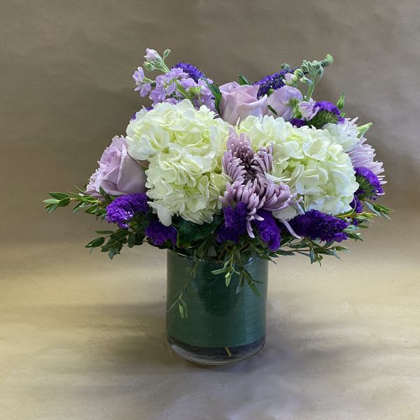 Lavender Frost - A lovely arrangement of luscious lavenders and purple nestled with white hydrangeas and placed within a leaf ribbon lined cylinder.