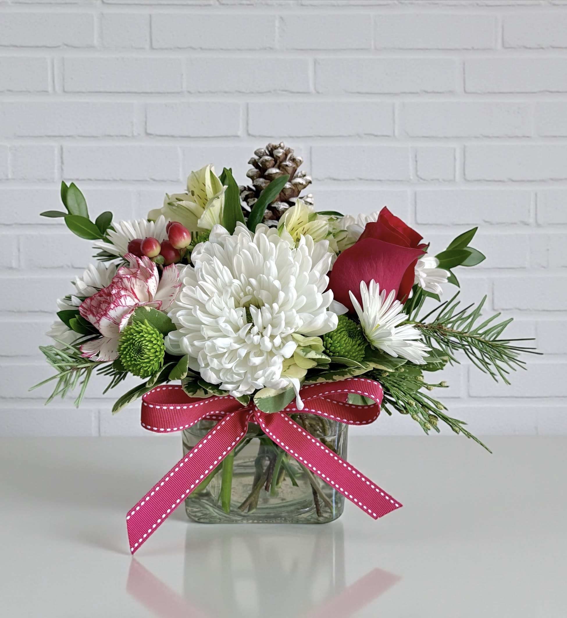 Small Holiday Cube - A 4” by 4 clear glass cube filled with winter greens, a white cremon, peppermint carnation, mums, alstroemeria and a red rose. 