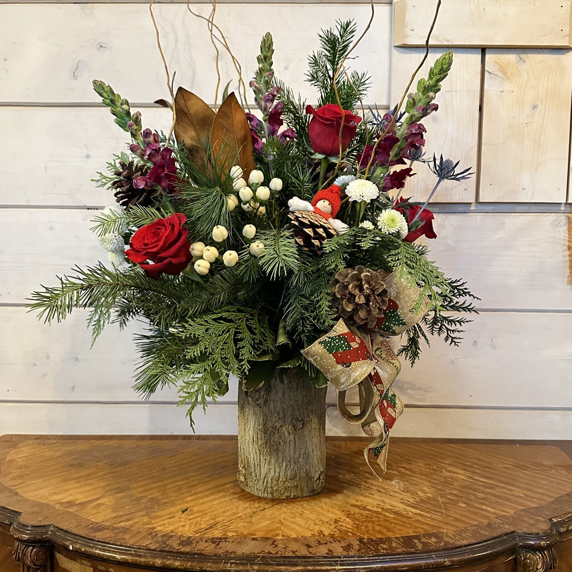 The Christmas Cabin Bouquet - Red roses, blue thistle, burgundy snapdragons, white button chrysanthemums, white hypericum berries, and a magnolia branch, arranged in a tall log pot with conifer greens, pinecones, a bow, curly willow, and a pinecone gnome. Approximately 20" high and 16" wide.
