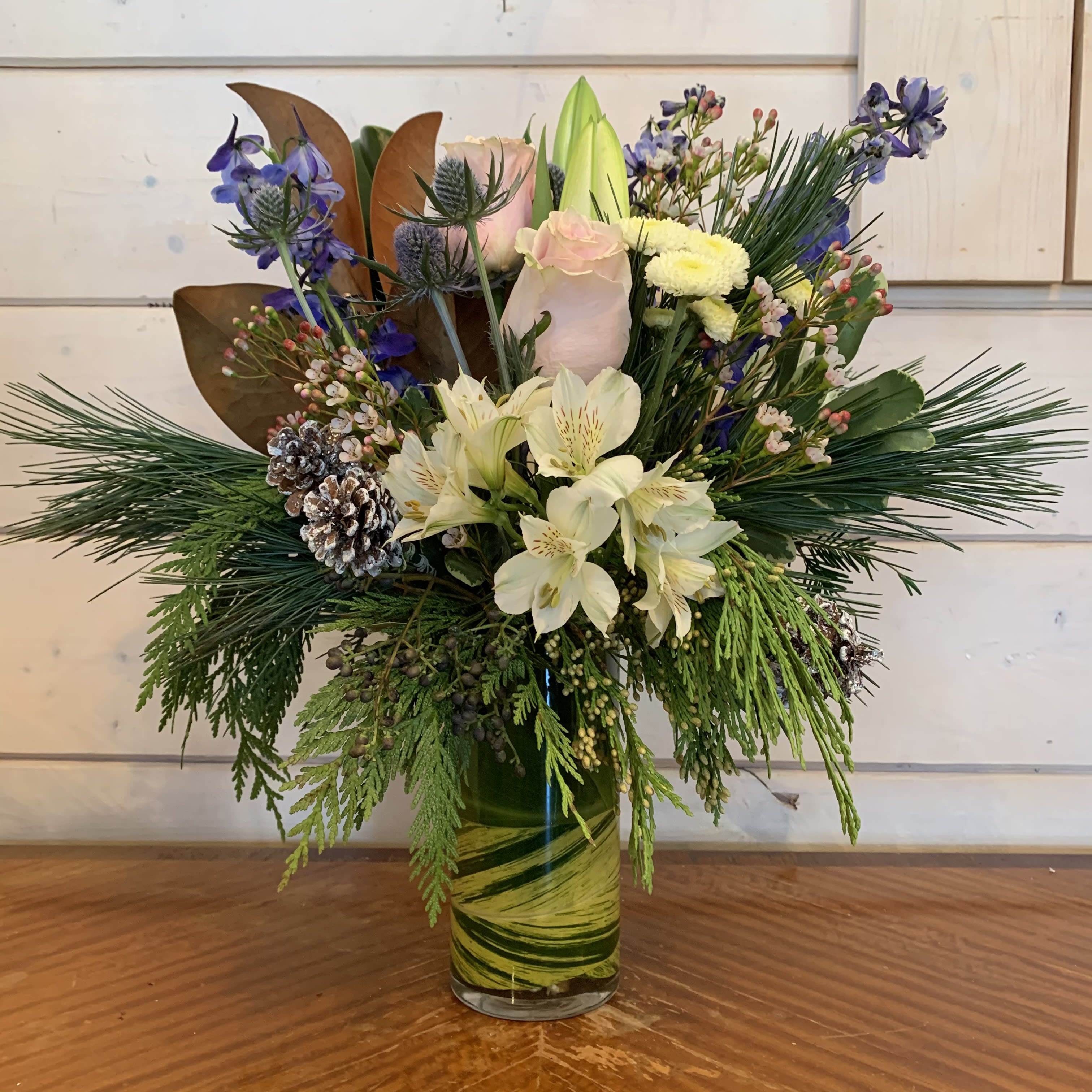Welcome Winter  - Pink mondail roses, white lily, blue delphinium, magnolia, white buttons, and white alstromeria arranged in a leaf lined 7in cylinder with blue thistle, white wax flower and white glitter pinecones. Approximatly 18in tall and 12in wide. 