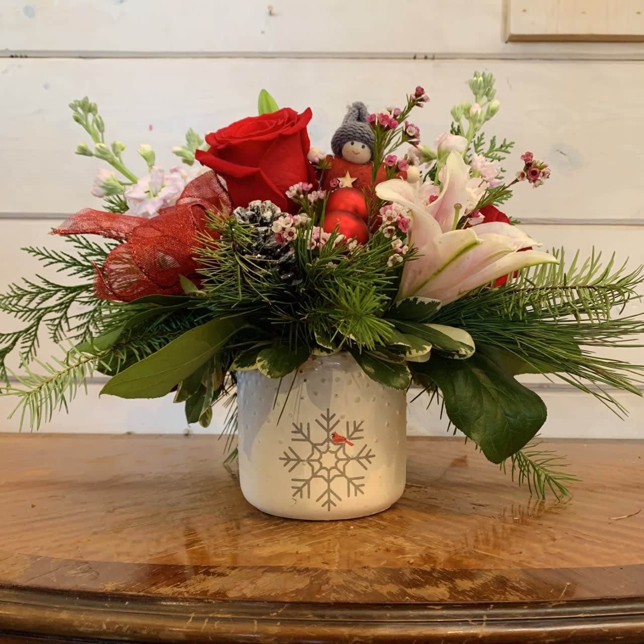 Winter Kisses Bouquet  - red roses, pale pink stock, stargazer lilies arranged in a small white ceramic pot with a snowflake and cardinal decal on the front. Seasonal winter evergreens, red bow, red ornament balls and snowy pinecones along with seasonal filler flowers. Small winter child ornament included. 