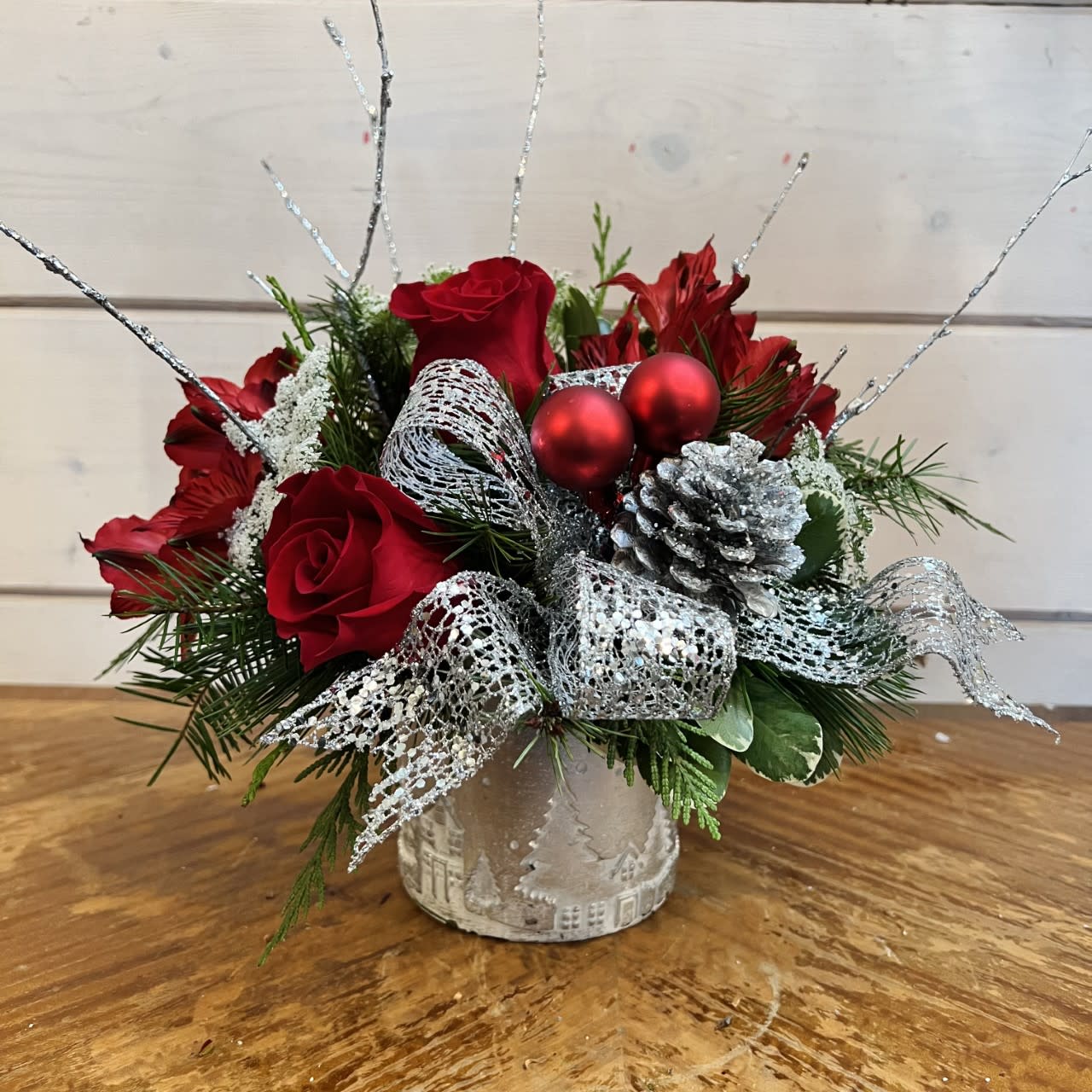 Silver Sleigh bells  - Red roses, red alstroemeria, queen ann lace and silver glitter branches arranged in a small silver ceramic city scape pot with seasonal evergreens, festive silver bow silver pinecones and red ornament balls. Approximately 10in tall and 10in wide. 