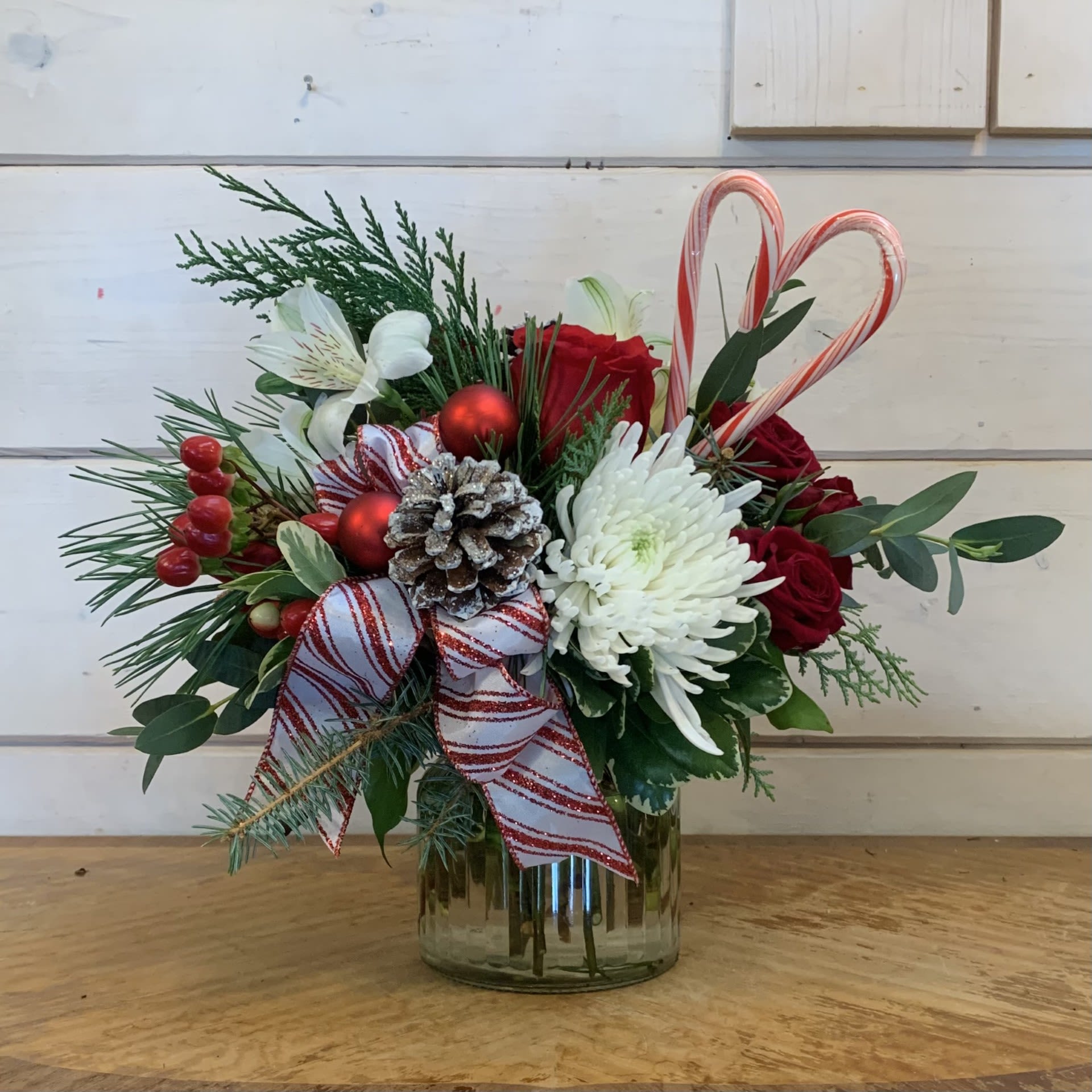The Peppermint Forest - Red roses, white cremon chrysanthemums, burgundy spray roses, red hypericum berries, and white alstroemeria arranged in a small glass ginger jar with red and white striped bow, glitter pinecones, red ornament balls, and 2 candy canes in the shape of a heart. Approximately 10&quot; tall and 10&quot; wide.