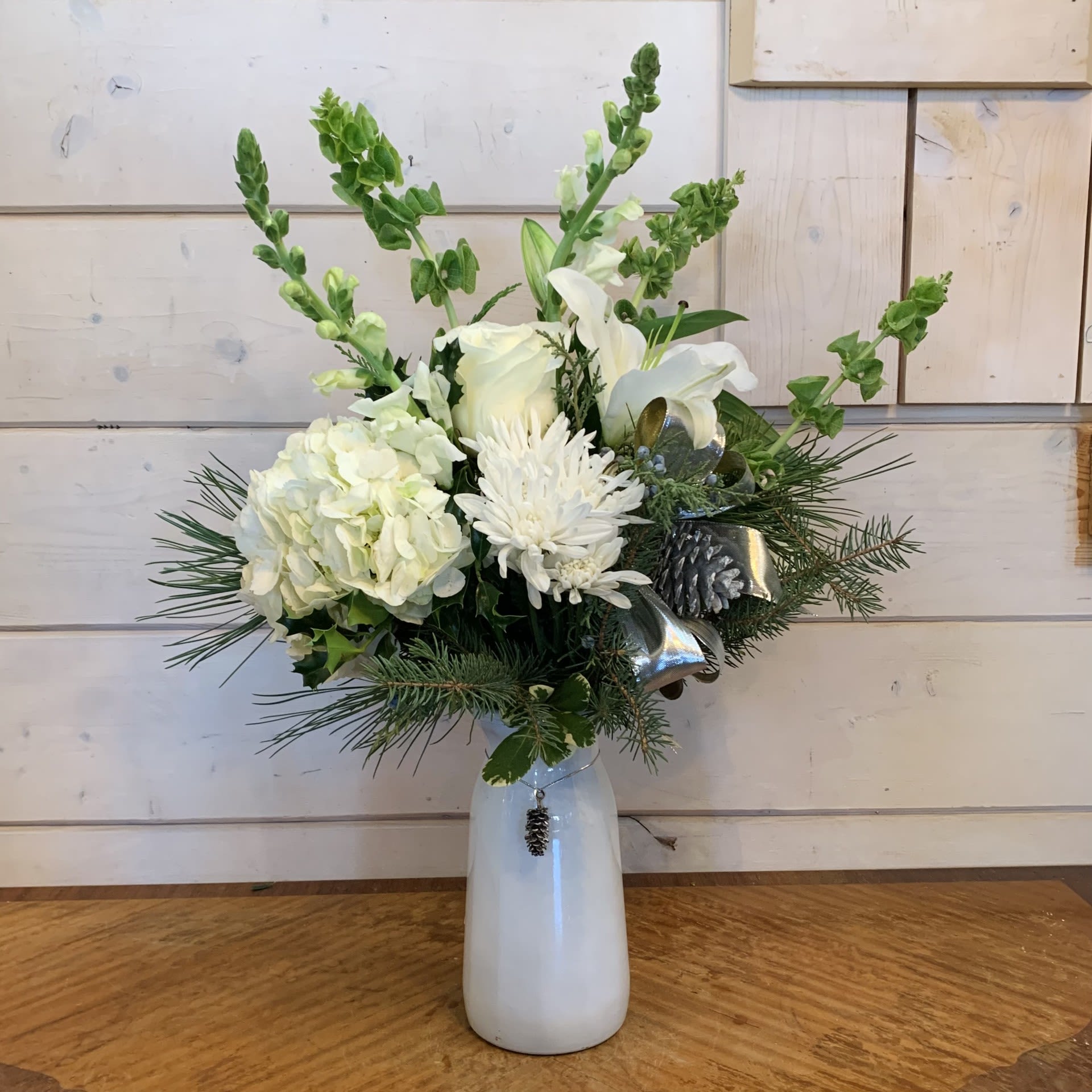 Winter Wonderland  - An elegant all white winter arrangement of white hydrangea, white lilies, white snapdragons, white roses, green bells of ireland and white chrysanthemums in a tall white vase with pinecone charm with silver bow and silver pinecones. Approximately 17&quot; tall by 11&quot; around.