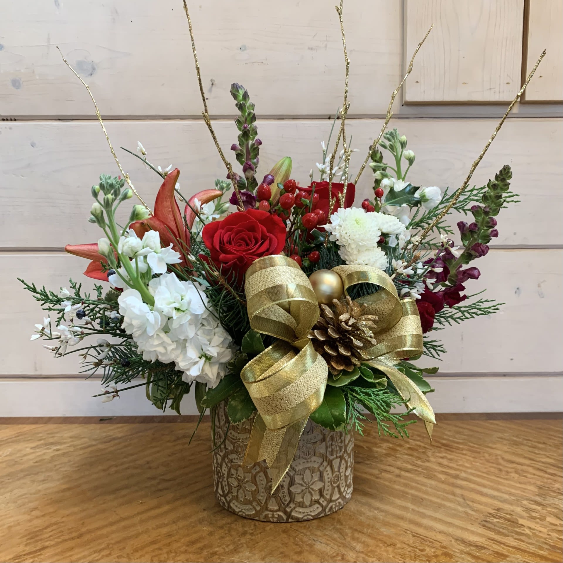 Yuletide Glow  - Red roses, burgundy lilies, and burgundy velvet snapdragons with fragrant white stock, red hypericum berries, and white crysanthemums in a gold scrolled pot with seasonal filler flowers, evergreen foliage, a gold bow, gold ornament balls, gold pinecones, and gold glitter branches. Approximately 10&quot; wide and 10&quot; tall.