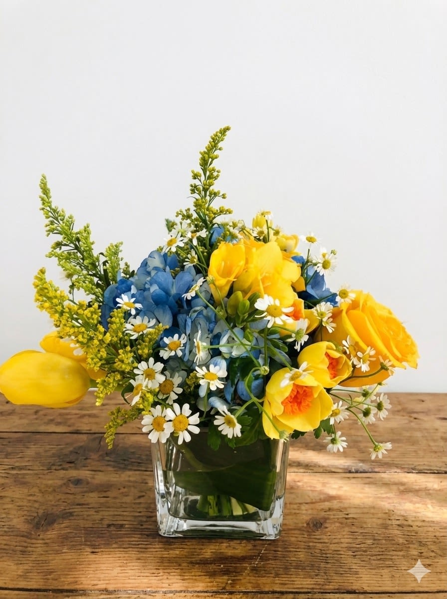 Spring in Cube - When we think of spring, this yellow and blue floral design comes to mind!  Cheerful chamomile flowers, yellow freesia, yellow tulips, yellow roses, blue hydrangea and yellow solidago wildflowers create the perfect spring vibe. You’ll be transported to warm spring days as soon as you see this flower arrangement! The design is presented in a 4&quot; clear glass cube vase. Standard size pictured. Larger sizes available. Flowers and vase will vary based on availability.