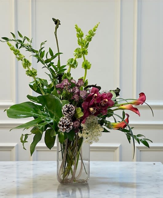 Orchids in Winterwood - A tall, sculptural composition that blends refined elegance with quiet winter textures. Sleek orchid blooms rise through natural branches dressed in soft moss, while subtle pine cones add an understated seasonal touch. Designed with a minimal, organic aesthetic, this piece brings a serene winter feel—elevated, modern, and effortlessly luxurious. Orchid color may vary based on availability.