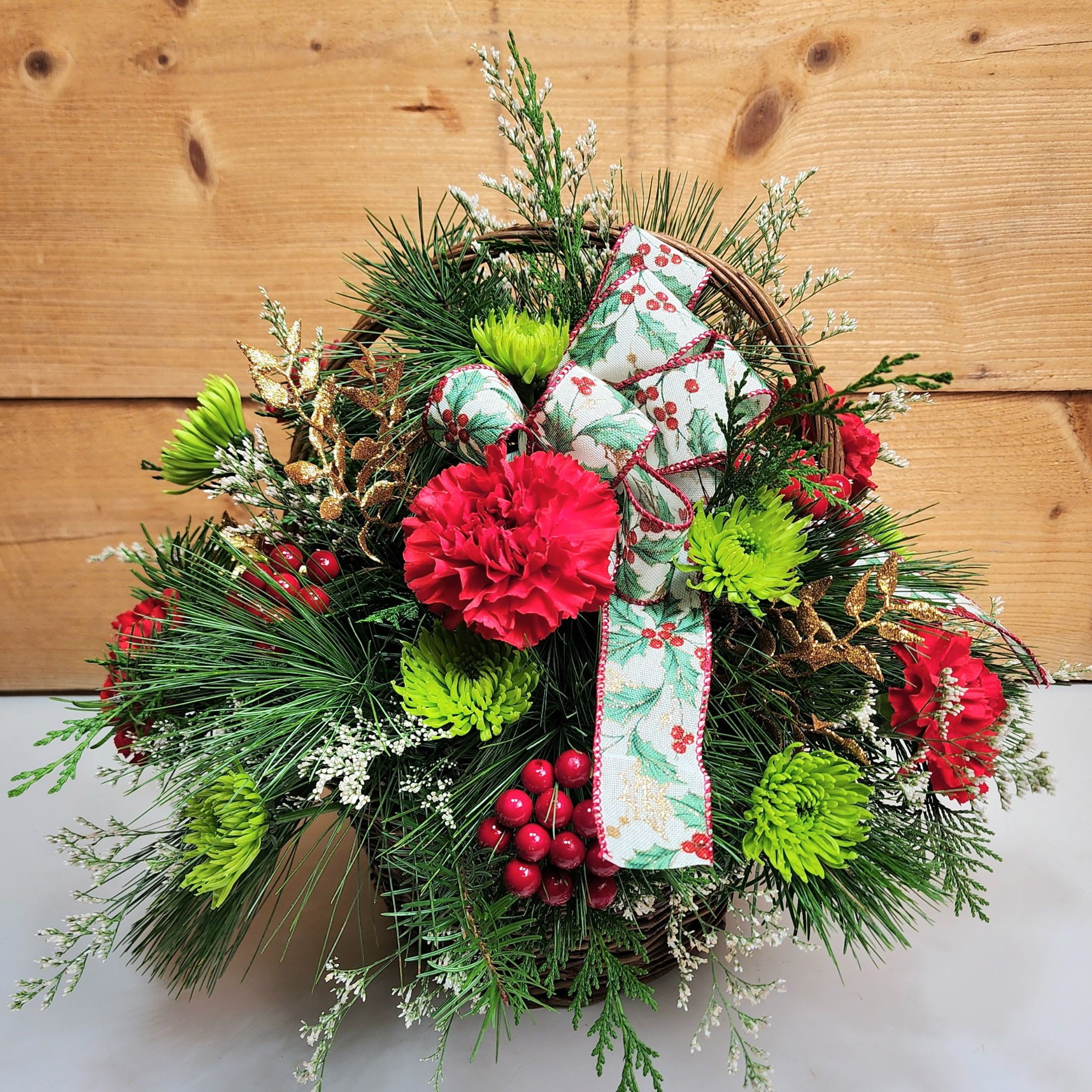 Holly Jolly Basket - Bring on the festivities with our Holly Jolly Basket! Carnations and pom poms are joined with faux berries and golden, glittery stems of leaves in this wonderful wicker basket, and the holly bow is the cherry on top to make this basket the perfect blend of traditional holiday decor and fun, fresh blooms!  Dimensions approx. L 16” x H 16”