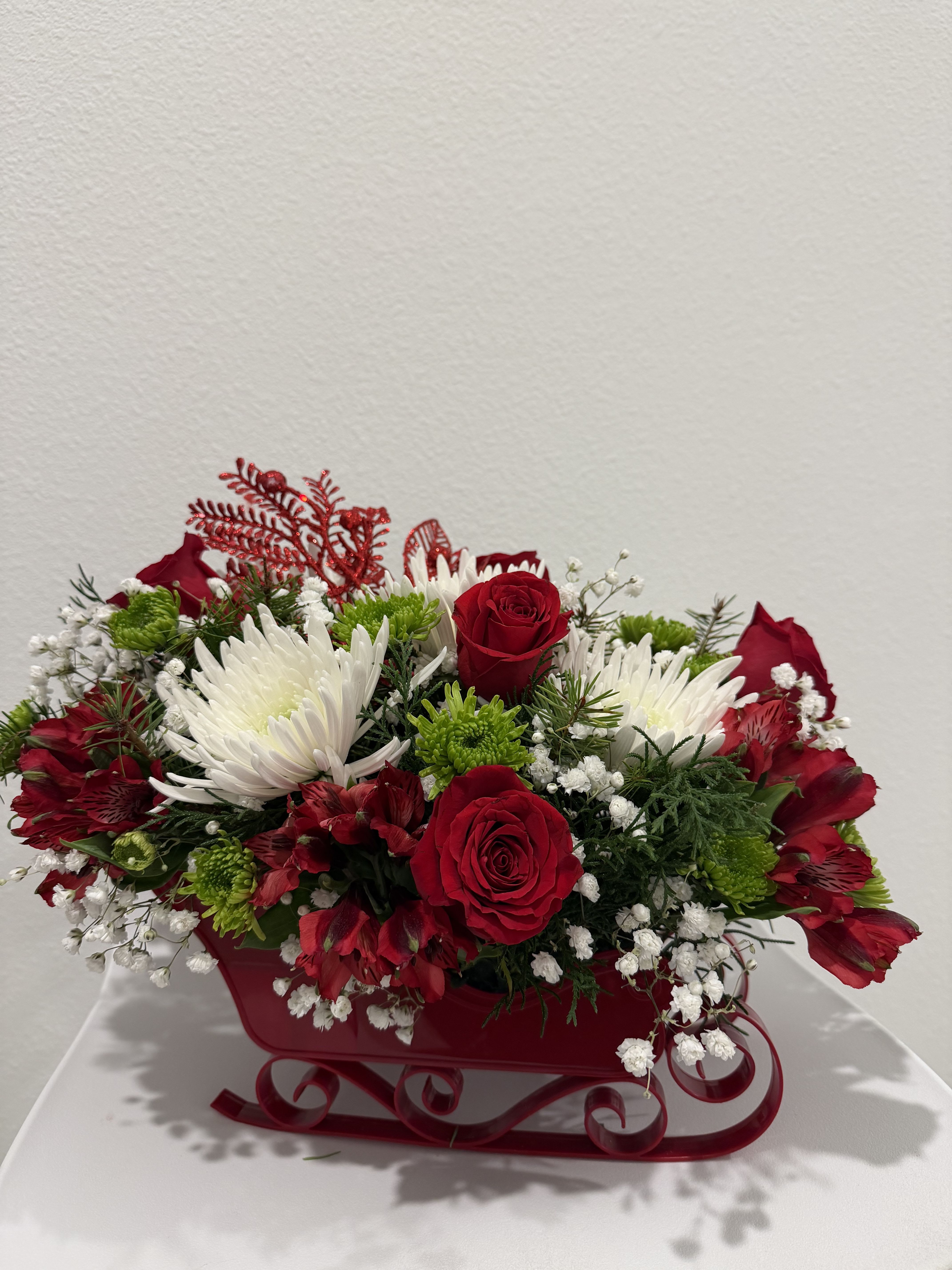 Holiday Sleigh Charm - A luxurious holiday arrangement nestled in a metal red sleigh, combining lush red roses, crisp white spider mums, delicate baby’s breath, and fresh greenery. Ideal for gifting or enhancing any Christmas décor with elegance and charm. 