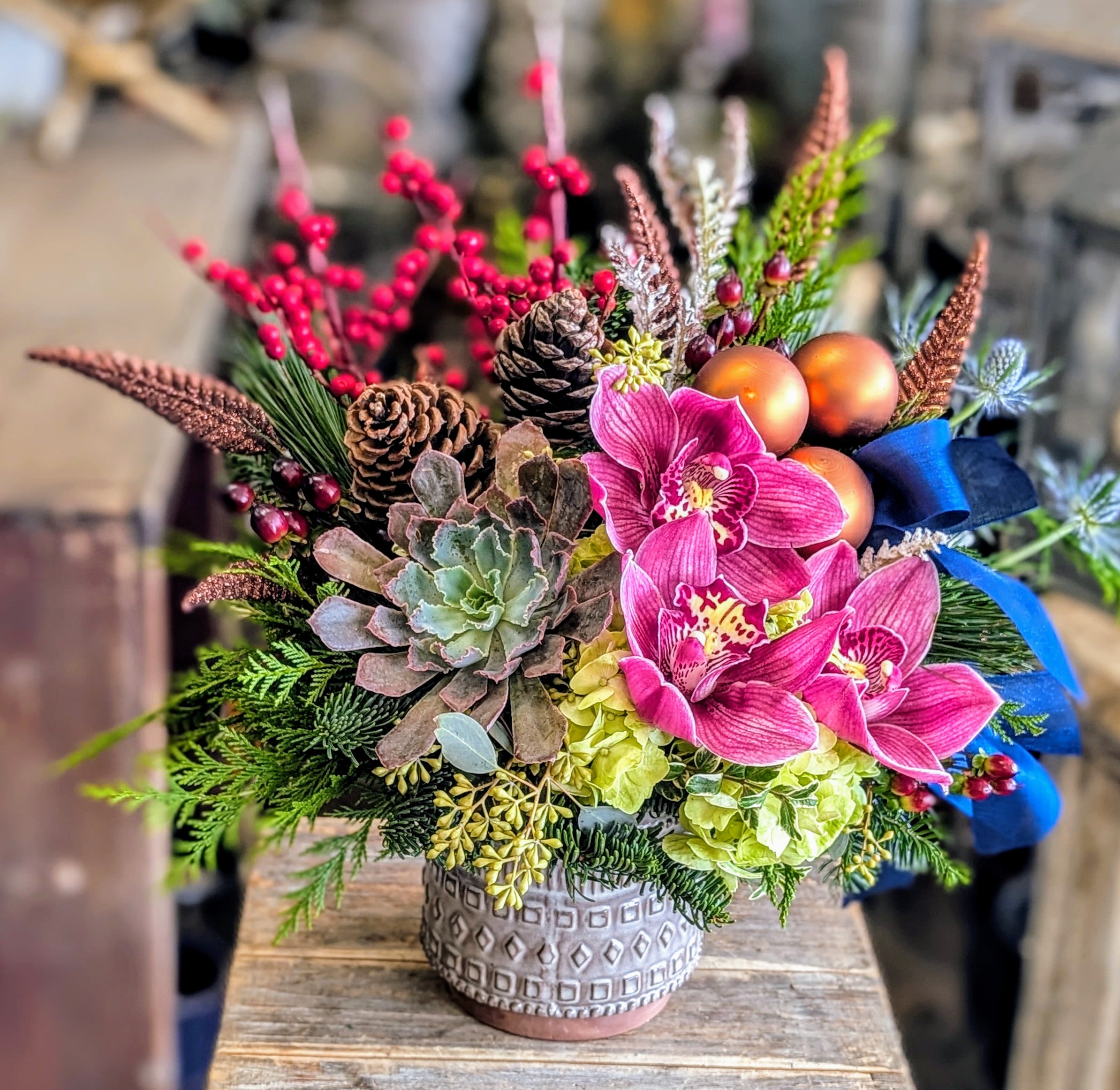 Nordic - Nordic – A Modern Winter Bloomscape  Minimal, modern, and beautifully bold, Nordic captures the essence of winter through rich textures and clean seasonal color. Handcrafted by Windmill Floral Expressions in Las Vegas, this arrangement blends luxe botanicals with cool-toned holiday accents—creating a contemporary winter statement piece that feels fresh, artistic, and effortlessly sophisticated.  Vibrant magenta cymbidium orchids take center stage, contrasted by plush green hydrangea and a full sculptural succulent rosette. Long-needle pine, cedar, and winter greenery add depth and movement, while pinecones and blue thistle bring in that crisp Nordic woodland texture. Copper ornaments and a sleek navy ribbon add a modern edge and polished holiday finish.  Perfect for homes with a clean, modern aesthetic, Nordic is a winter arrangement designed for clients who appreciate artistry, color, and winter freshness without traditional heaviness.  Perfect For:  • Winter décor with a modern, Scandinavian-inspired feel • Holiday gifts in Las Vegas &amp; Henderson • Succulent lovers seeking seasonal flair • Minimal or contemporary interior styles