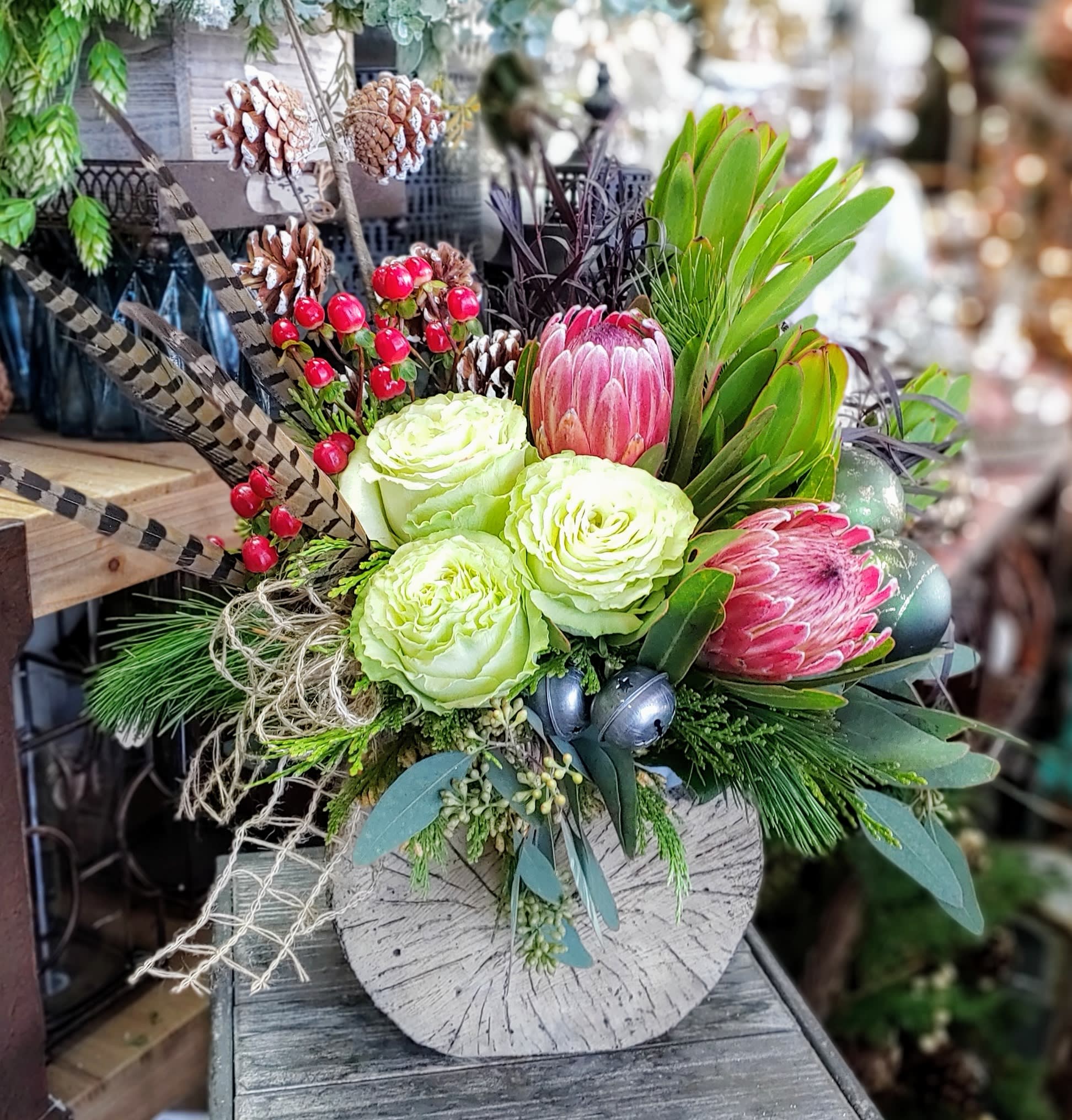 Protea Winter - Protea Winter – A Sculptural Holiday Arrangement with Rustic Elegance  Bold, textural, and beautifully unexpected, Protea Winter showcases the natural artistry of the season. Handcrafted by Windmill Floral Expressions in Las Vegas, this arrangement blends striking blooms with winter woodland textures to create a holiday piece that feels both modern and organic.  Soft green garden roses pair effortlessly with rosy pink protea, offering a vivid contrast of color and form. Sprays of winter berries, pinecones, and seasonal greens add movement and freshness, while pheasant feathers and natural jute fibers introduce a rustic, earthy charm. Metallic ornaments tucked between the foliage add a subtle holiday shimmer, enhancing the sculptural beauty of the protea.  Designed in a textured stone-inspired vessel, Protea Winter brings together wild botanical elements and modern winter style—perfect for clients seeking something unique, artistic, and full of seasonal personality.  Perfect For:  • Rustic chic winter décor • Unique holiday gifts in Las Vegas &amp; Henderson • Protea lovers and fans of sculptural blooms • Homes with natural, modern, or eclectic style