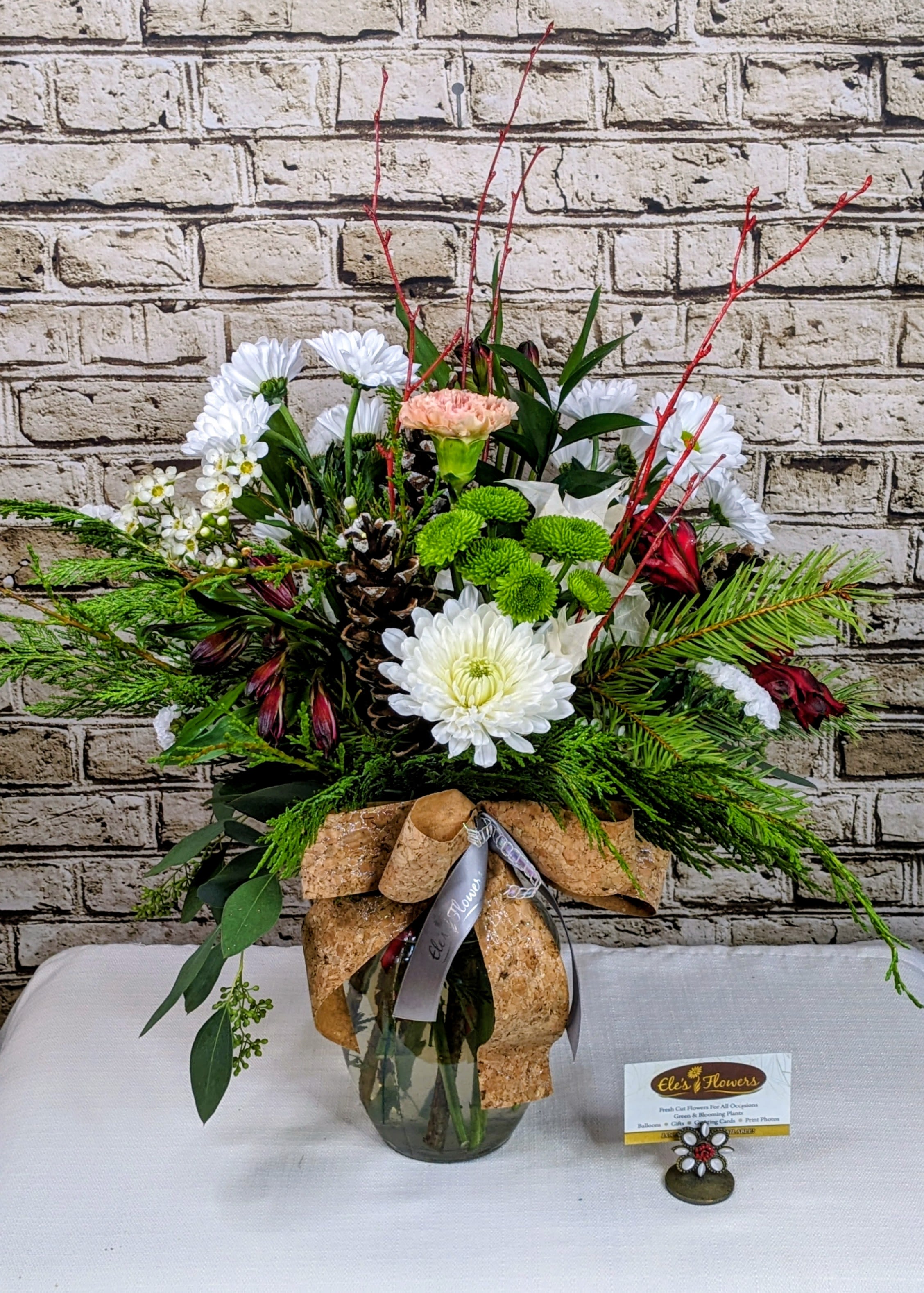 Winter Woodland Glow  - Bring the magic of a snowy forest indoors with our Winter Woodland Glow arrangement. This design blends fresh evergreens, crisp white blooms, and pops of rich holiday color to create a lush, nature-inspired display that feels both cozy and elegant.  Accented with frosted pinecones, vibrant green button blooms, and seasonal red branches, this arrangement captures the charm of a winter stroll through the woods. A soft peach blossom adds a gentle touch of warmth, while the cork-inspired bow gives the entire piece a rustic, handcrafted look—perfect for holiday gifting or elevating your own seasonal décor.  Ideal for Christmas celebrations, winter birthdays, office surprises, or simply bringing a touch of festive cheer to any space, Winter Woodland Glow is a fresh, welcoming statement of the season.  Approximate Dimensions: 22&quot;H x 18&quot;W