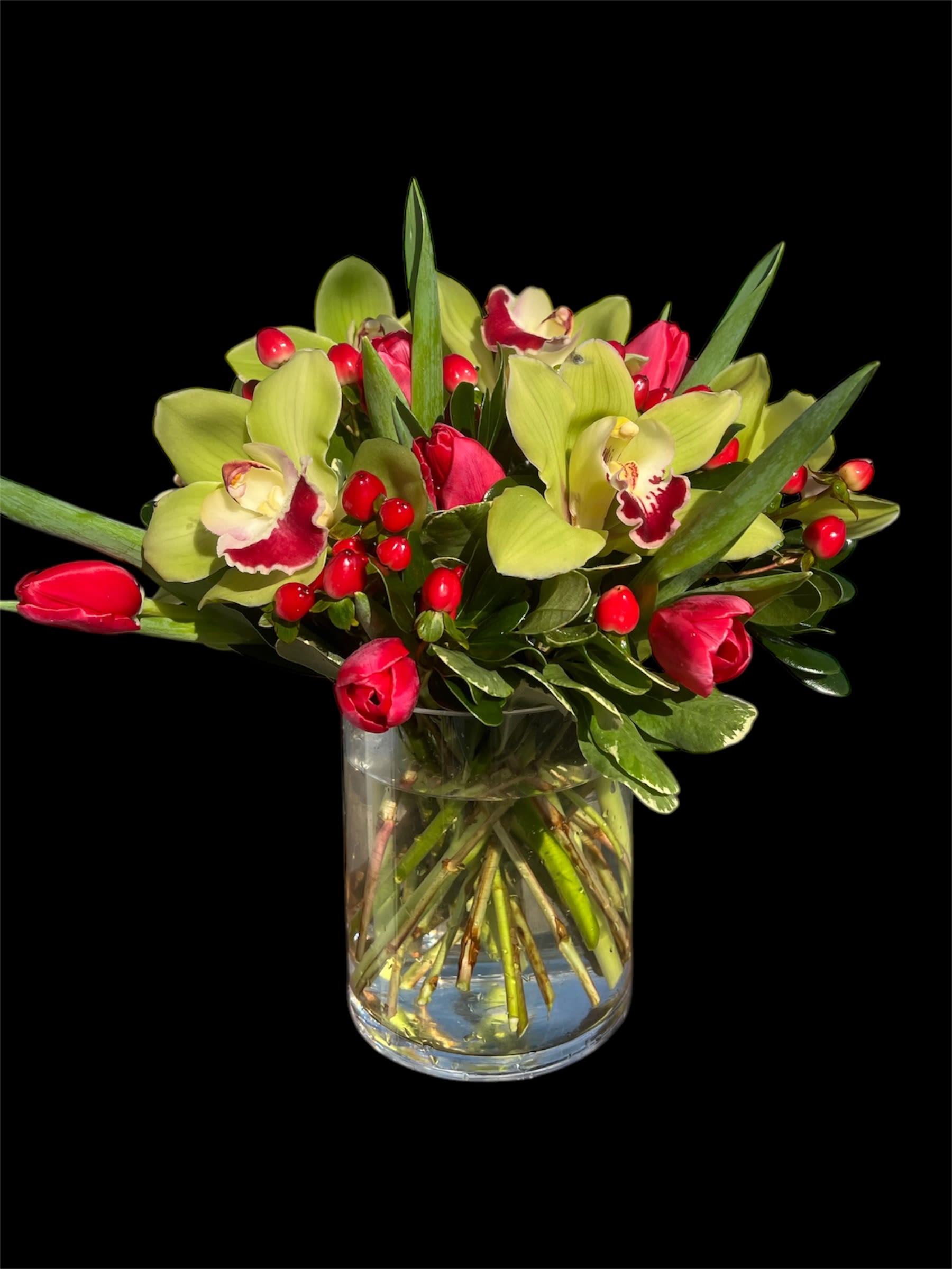 Sweetheart bqt - A vibrant and captivating arrangement featuring lush green cymbidium orchids, rich red hypericum berries, and striking red tulips. Nestled in a sleek cylinder glass vase, this combination creates a dynamic and eye-catching display, perfect for adding a touch of elegance and warmth to any setting.