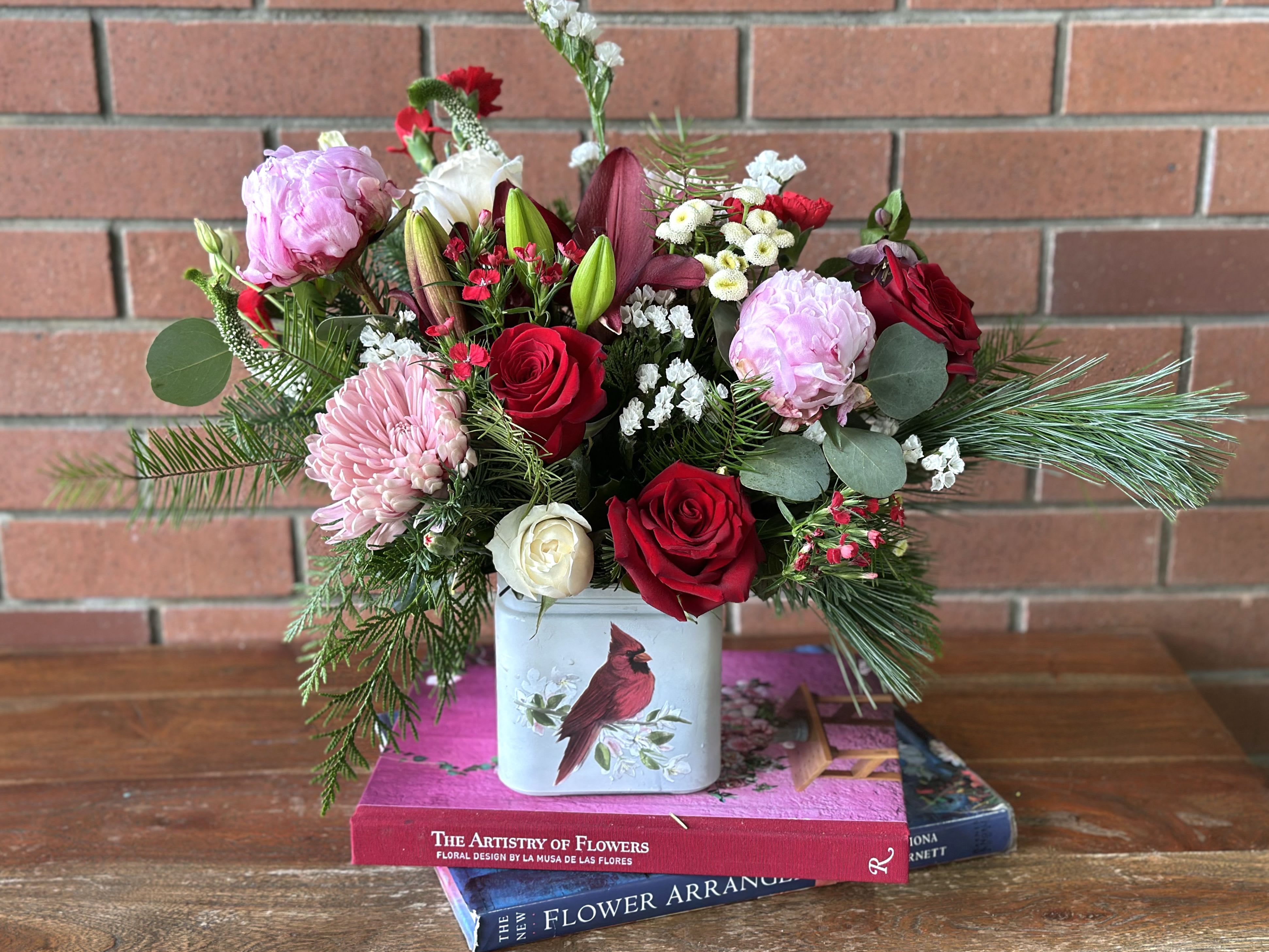 Cardinal’s Song Christmas Bouquet  - The &quot;Cardinal's Song Christmas Bouquet&quot; by Venetian Flowers a stunning floral arrangement that captures the essence of the holiday season. It features a beautiful square vase that serves as a perfect backdrop for a vibrant red cardinal perched gracefully among a rich tapestry of mixed evergreens. The arrangement bursts with color, showcasing striking red lilies, lush peonies, and romantic red roses, all harmoniously combined with cheerful mums and delicate red mini carnations. To add a touch of elegance, spray roses offer a charming accent, while white filler flowers provide a lovely contrast, enhancing the overall beauty of the bouquet. This arrangement not only celebrates the beauty of nature but also evokes the warmth and joy of Christmas, making it a perfect centerpiece for festive gatherings