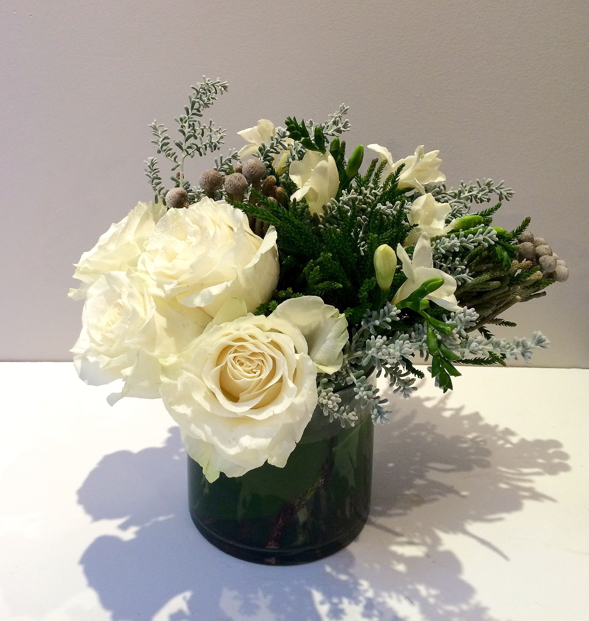 Snowy Day - A representation of the winter wonderland, composed of crystal white garden roses, white fragrant freesias, winter brunias, green pines and eucalyptus seeds in round glass container.  Size: 13"w x 10"h x 7"d
