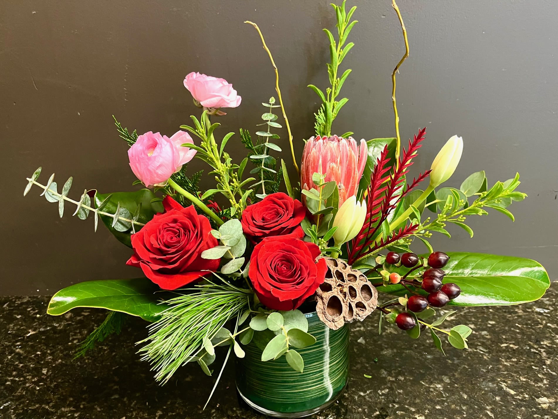 Holiday Prairie - festive greens are collected in a glass container accented with red roses, protea, ranunculus and tulips 
