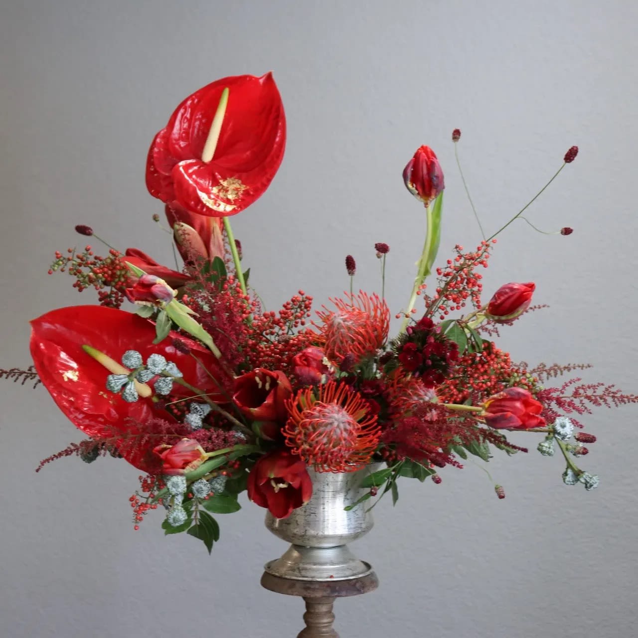 Merry Berry Bliss - This stunning arrangement captures the festive spirit of the holiday season with its rich crimson hues and elegant composition.  The bold red anthuriums, tulips, and pincushion proteas evoke the warmth and joy of the holidays, while subtle silver eucalyptus adds a frosted touch, reminiscent of winter magic.  Perfect as a centerpiece for holiday gatherings or as a luxurious gift, this design combines modern artistry with the timeless beauty of the season, creating a festive statement of elegance and celebration.   ( Please note that flowers may vary depending on season and availability, but our expert florists will always craft each arrangement with the same attention to detail and quality. Any substitutions will be of equal or greater value and will not affect the overall beauty of the arrangement. )