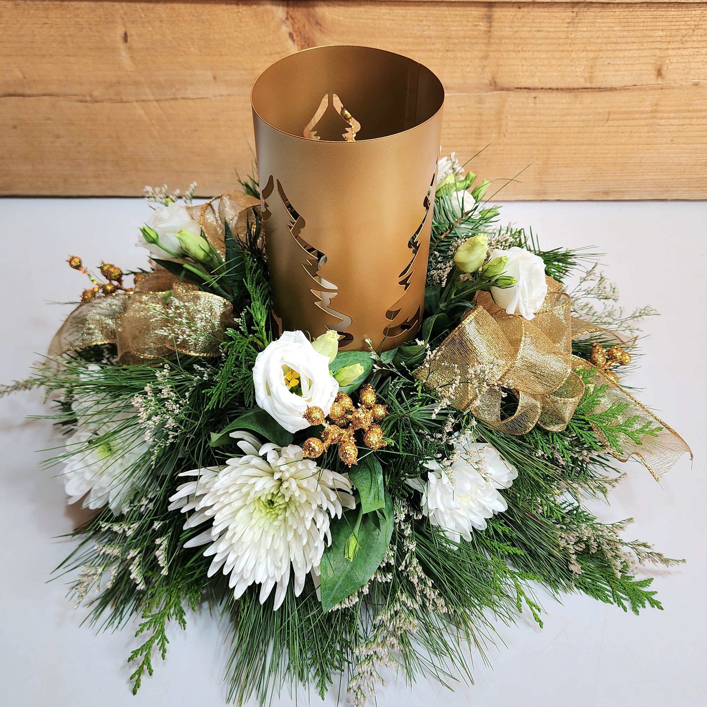 Golden Evergreen Centerpiece - Feel the warmth of holiday delight with our Golden Evergreen Centerpiece! This design features a golden lantern, with the shape of pine trees allowing for illumination from the candle inside of it. Along with the lantern, this arrangement comes with sophisticated white blooms like lisianthus and fuji mums, as well as golden berry accents to create a shining centerpiece for your holidays.  Dimensions approx. L 16&quot; x H 11&quot;