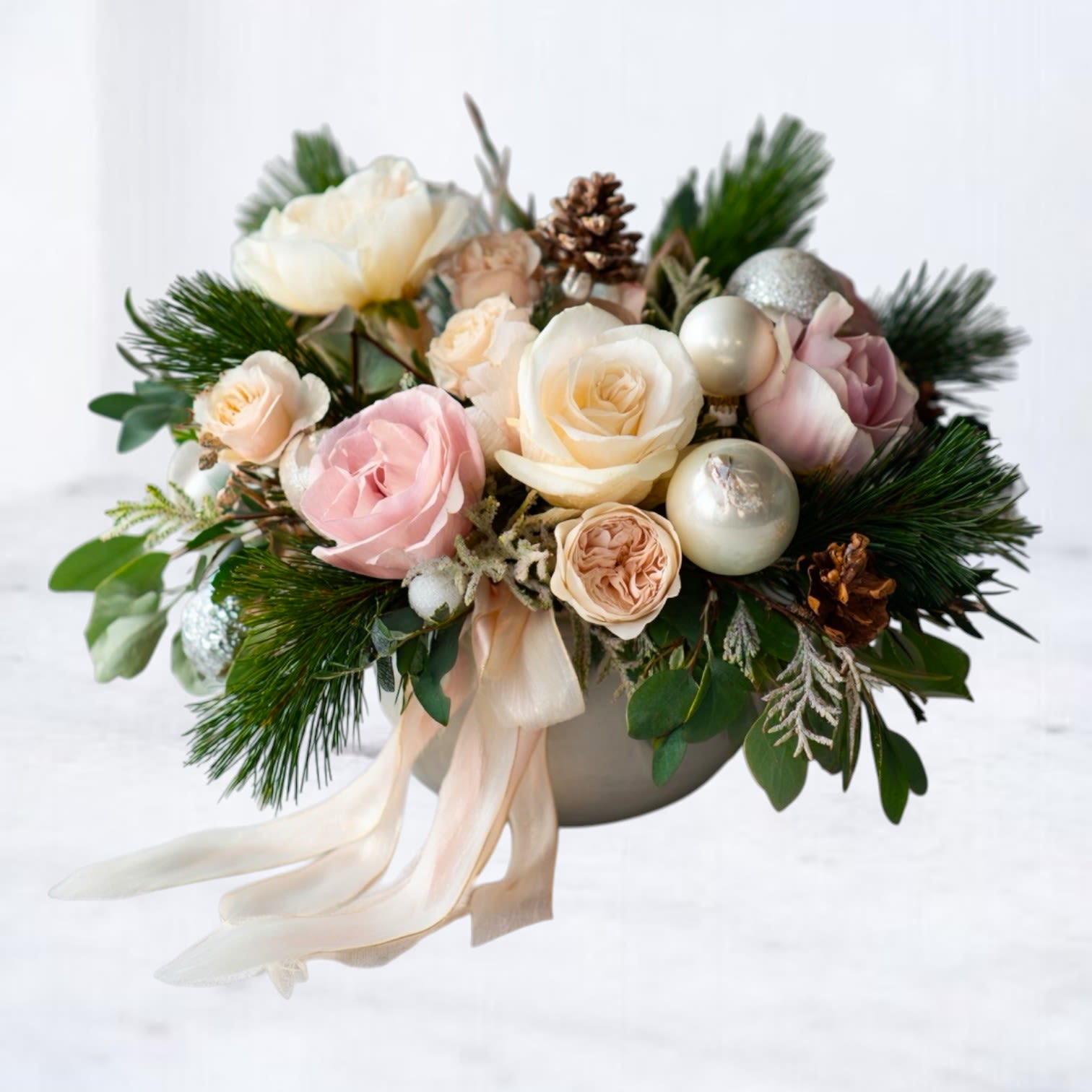 Pearl Petaline - Introducing the Pearl Petaline Arrangement — a soft, romantic winter design crafted with pastel roses, creamy garden blooms, and lush seasonal evergreens. Delicate pearl-toned ornaments and natural pinecones add dimension and glow, while airy textures and a flowing ribbon create a gentle, dreamlike finish.  Perfect for gifting, holiday entertaining, or adding a warm seasonal touch to your home, this arrangement blends elegance with cozy winter charm. Every piece is handcrafted at Whiskey Wallflower with premium flowers, fresh greenery, and thoughtful artistry.  Available for same-day flower delivery in Magnolia, The Woodlands, Montgomery, Conroe, and surrounding areas.  Substitution Disclaimer: Seasonal blooms and accents may vary based on availability. Any necessary substitutions will reflect the same palette, style, and value.