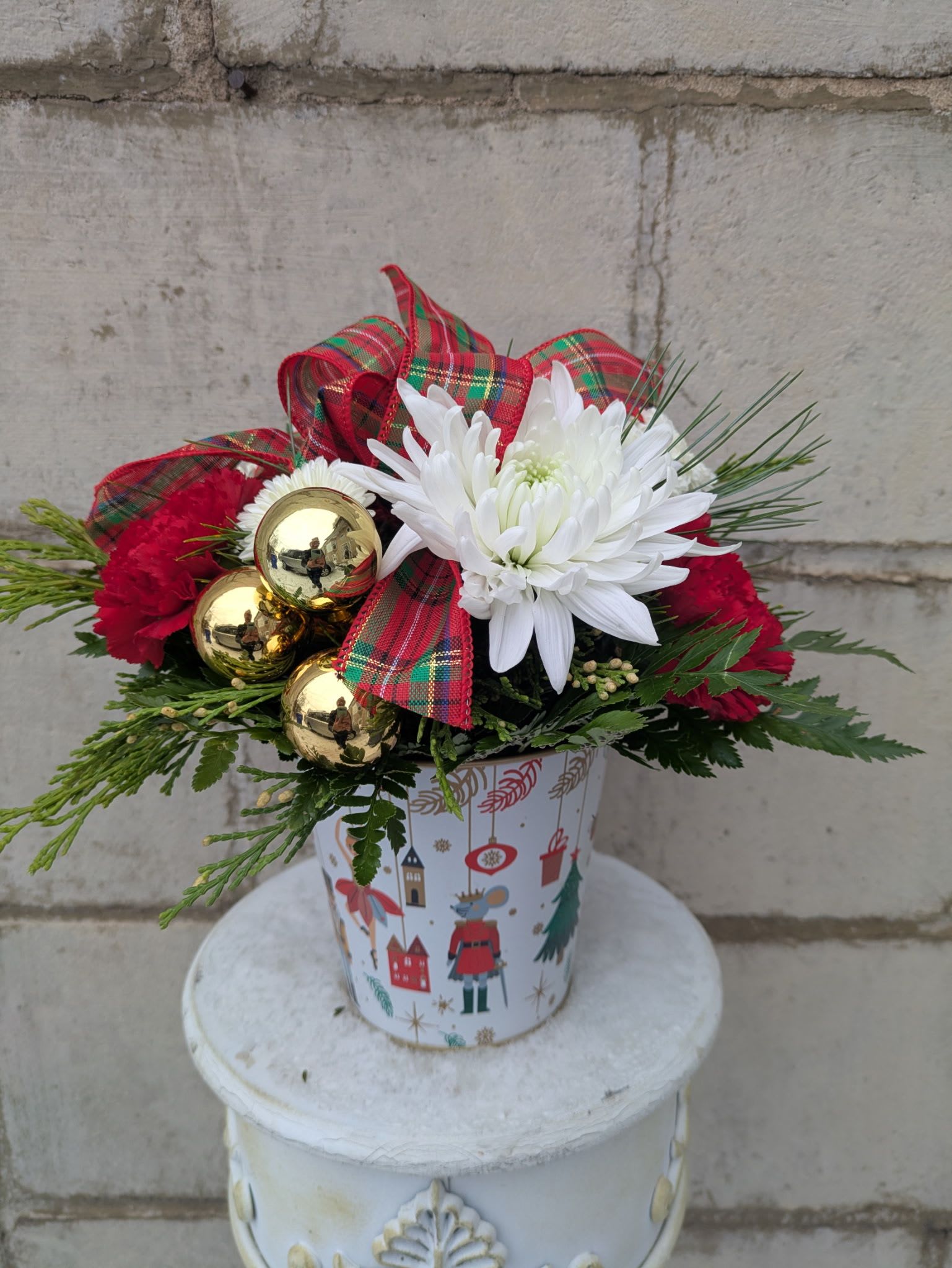 Holiday March  - Step into the magic of the season with Holiday March, a charming Christmas arrangement presented in an ornate nutcracker-themed container. Bursting with festive cheer, this design pairs crisp white Fuji mums, snowy white button poms, and vibrant red carnations for a classic holiday palette. Gleaming gold baubles add a touch of luxe, while a timeless plaid ribbon ties everything together with cozy seasonal warmth.  Perfect for gifting, decorating, or adding a whimsical touch to your holiday home, Holiday March brings the spirit of the Nutcracker to life—one joyful bloom at a time.  All flowers, floral colors, and containers are subject to change due to availability.